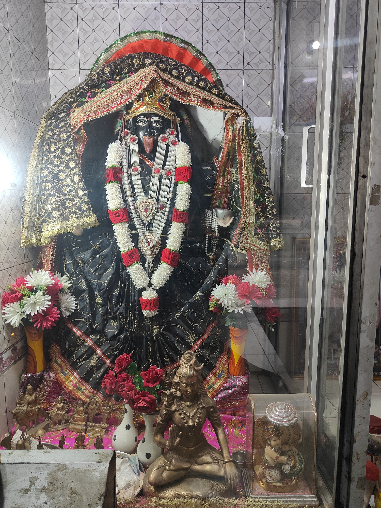 A full shot of a shrine to Kali, the Hindu goddess, is presented. The shrine is located behind a glass partition and is adorned with flowers and other offerings. The statue of Kali is black, with a stern expression and her tongue sticking out. She is decorated with garlands of white and red flowers and several necklaces. A crown sits atop her head. Two smaller statues, one of Shiva in a meditative pose and another of Krishna and Radha, are visible at the base of the shrine. The shrine is situated in a room with tiled walls. The overall setting suggests a religious space in Gurgaon, India.
