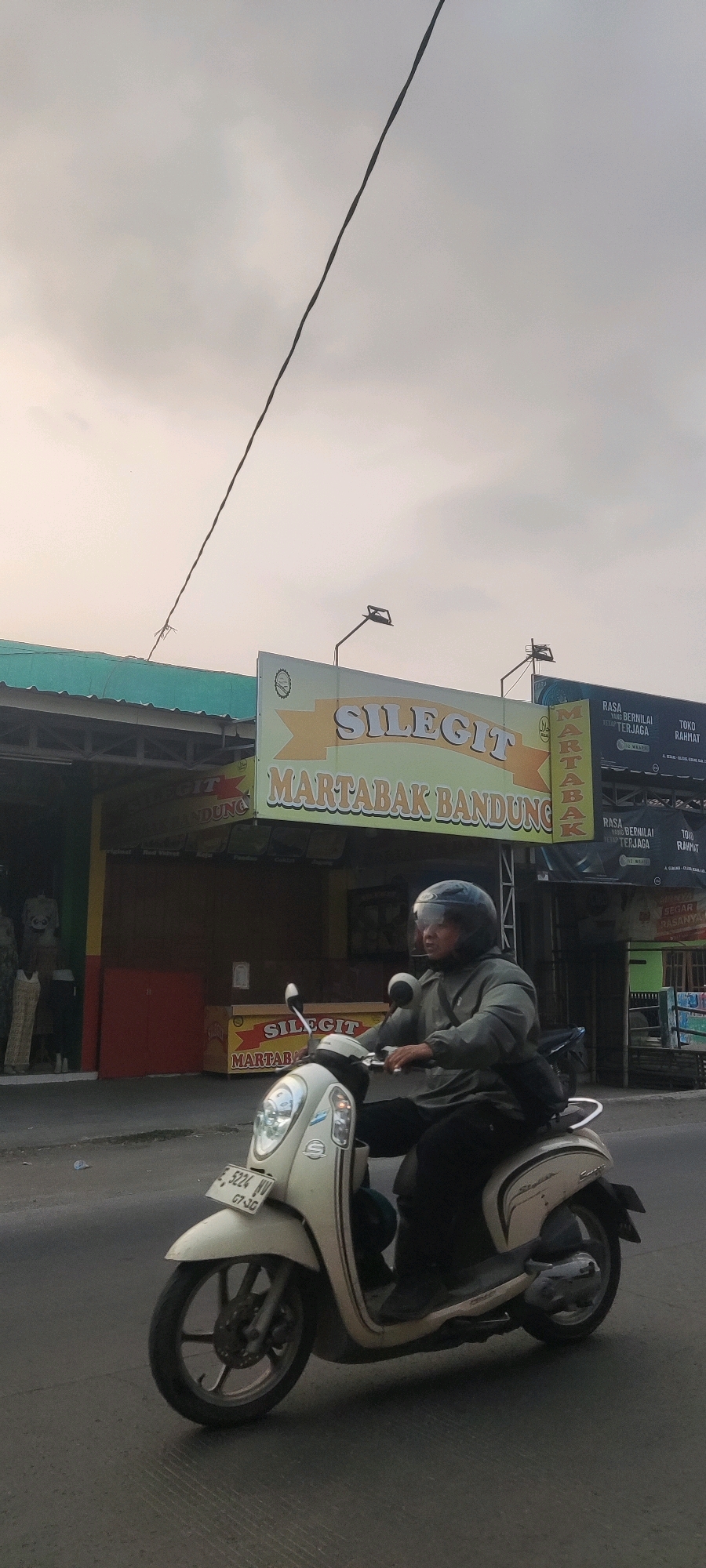 A person wearing a helmet and a grey jacket rides a cream-colored scooter on a paved road. The scooter has a license plate that reads "E 5224 WV GT-JG". Behind the scooter, a building with a green roof is visible, featuring signs with text in Indonesian. One sign, with a yellow background and a ribbon design, reads "SILEGIT MARTABAK BANDUNG". Another sign to the right has black text and a red background. The sky is overcast and light grey. The scene appears to be in an urban or suburban area, likely in Gebang, Indonesia, given the Indonesian text on the signs.