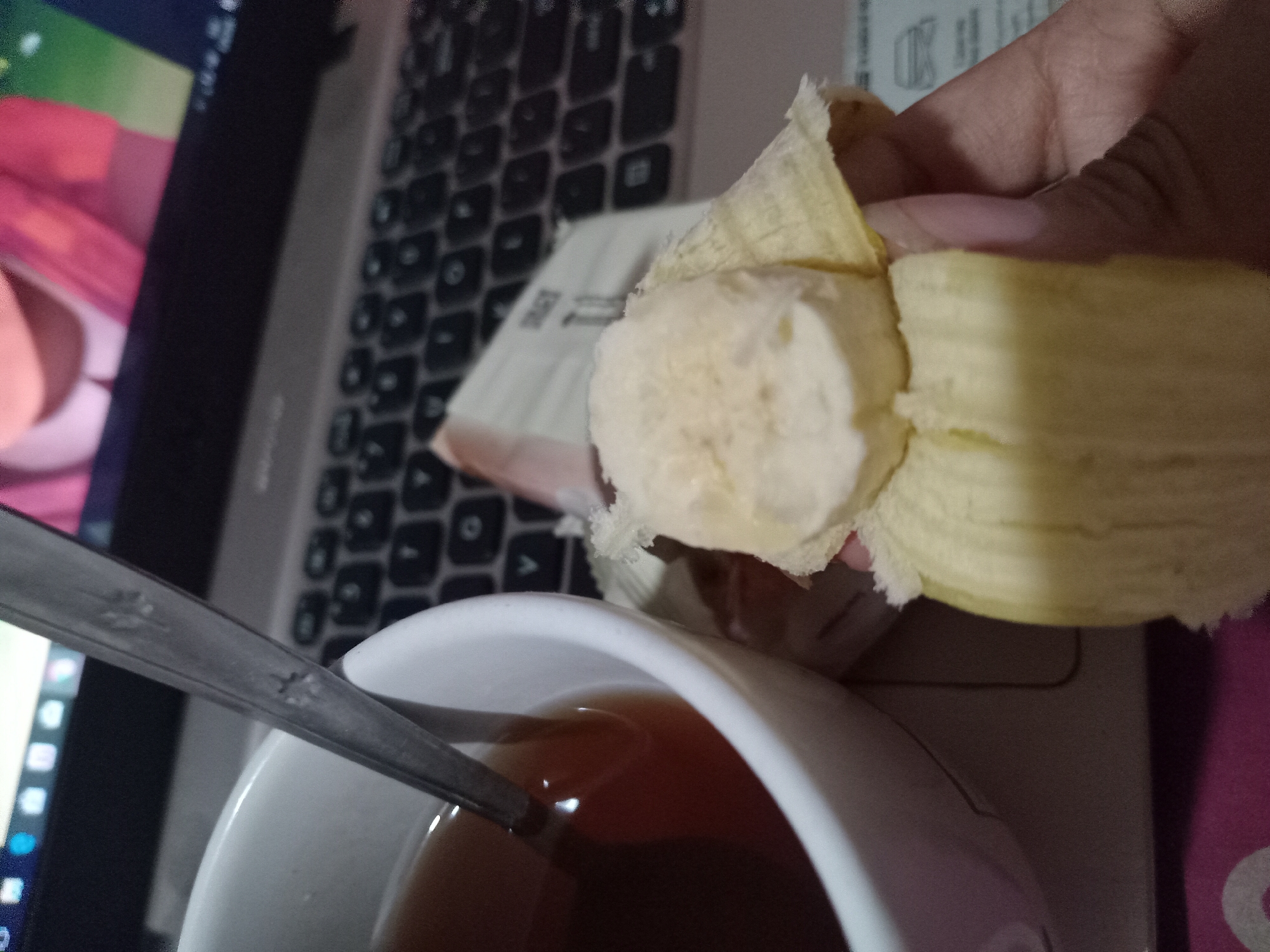 A close-up, eye-level shot shows a hand holding a peeled banana over a white mug of tea. A metal spoon rests in the mug, and the handle is visible to the left of the frame. A laptop keyboard is visible in the background, with a screen displaying a colorful image, possibly of an animated character. The scene appears to be indoors, with a pink and purple patterned surface visible at the bottom of the frame. No specific location in Claket, Indonesia, can be determined from the image alone.