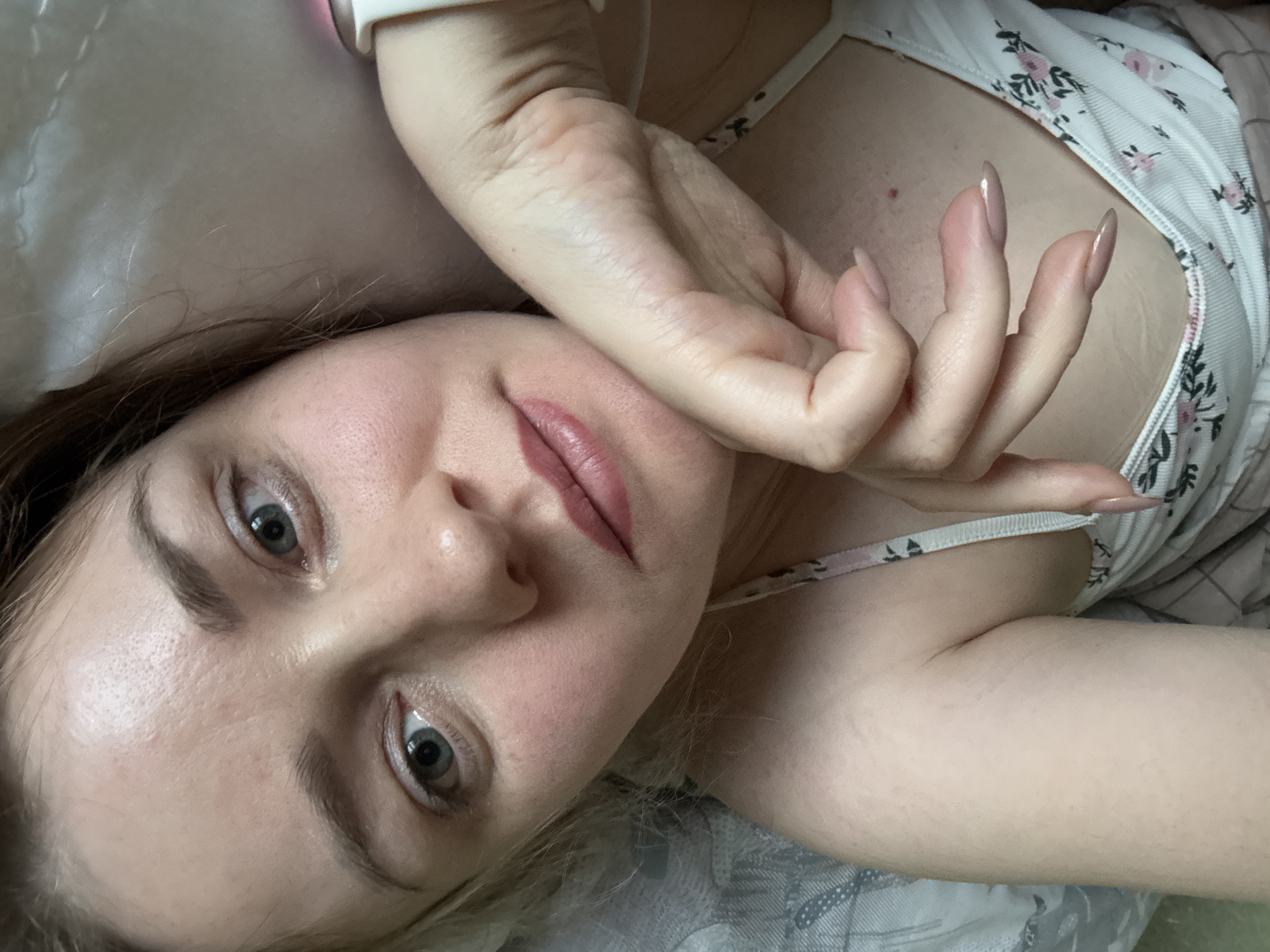 The media file captures a close-up portrait of a young woman lying down indoors, likely in a bedroom setting in Chaikovsky, Russia. She is positioned with her head slightly tilted, looking directly at the camera with an open, neutral to subtly engaging expression.
The primary subject is the woman, who appears to have fair skin, light blue or grey eyes, and medium brown hair. Her eyebrows are naturally defined, and her lips are adorned with a soft, natural pink lipstick. Her left hand is raised and positioned near her face, with long, almond-shaped nails painted in a light, neutral color. On her left wrist, she wears a light-colored wristband, possibly a smartwatch. She is dressed in a light-colored camisole or top featuring a delicate floral pattern of small pink flowers and green foliage. A small reddish spot is visible on her upper chest.
The background consists of soft, light-colored fabric, indicative of bedding or a pillow, which is slightly out of focus, emphasizing her face. The lighting is soft and natural, suggesting it is daytime. The overall scene conveys a relaxed and casual atmosphere. There is no visible text in the image.