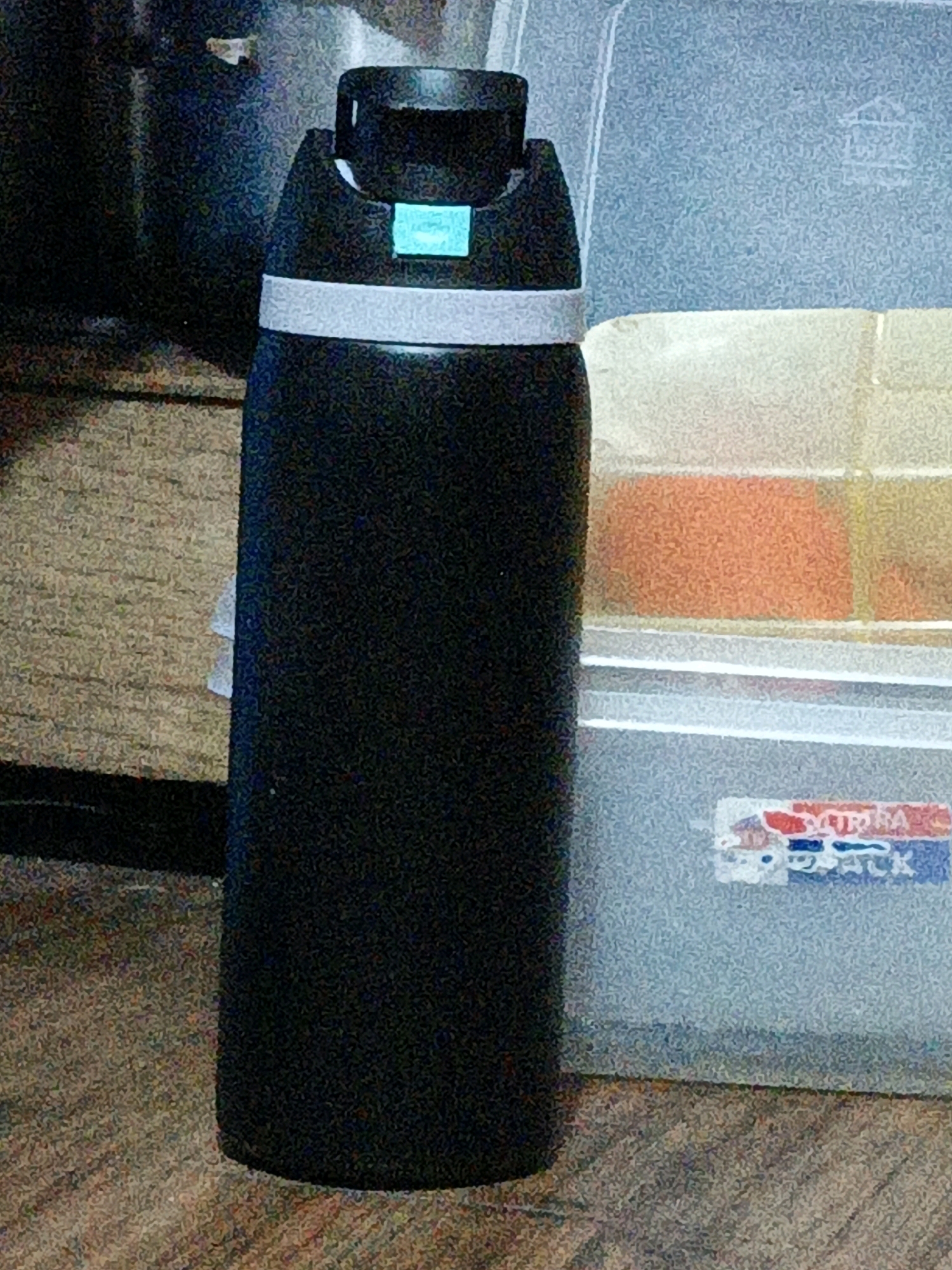 A black thermal flask with a light blue accent on its lid and a white band around its neck stands upright on a patterned floor, likely carpeted. Behind the flask, a translucent plastic container with an orange-colored object inside is visible. A portion of a sticker with red, white, and blue lettering is attached to the front of this container. The background is dark and out of focus. The image was taken indoors, in a residential setting in West Jakarta, Indonesia.