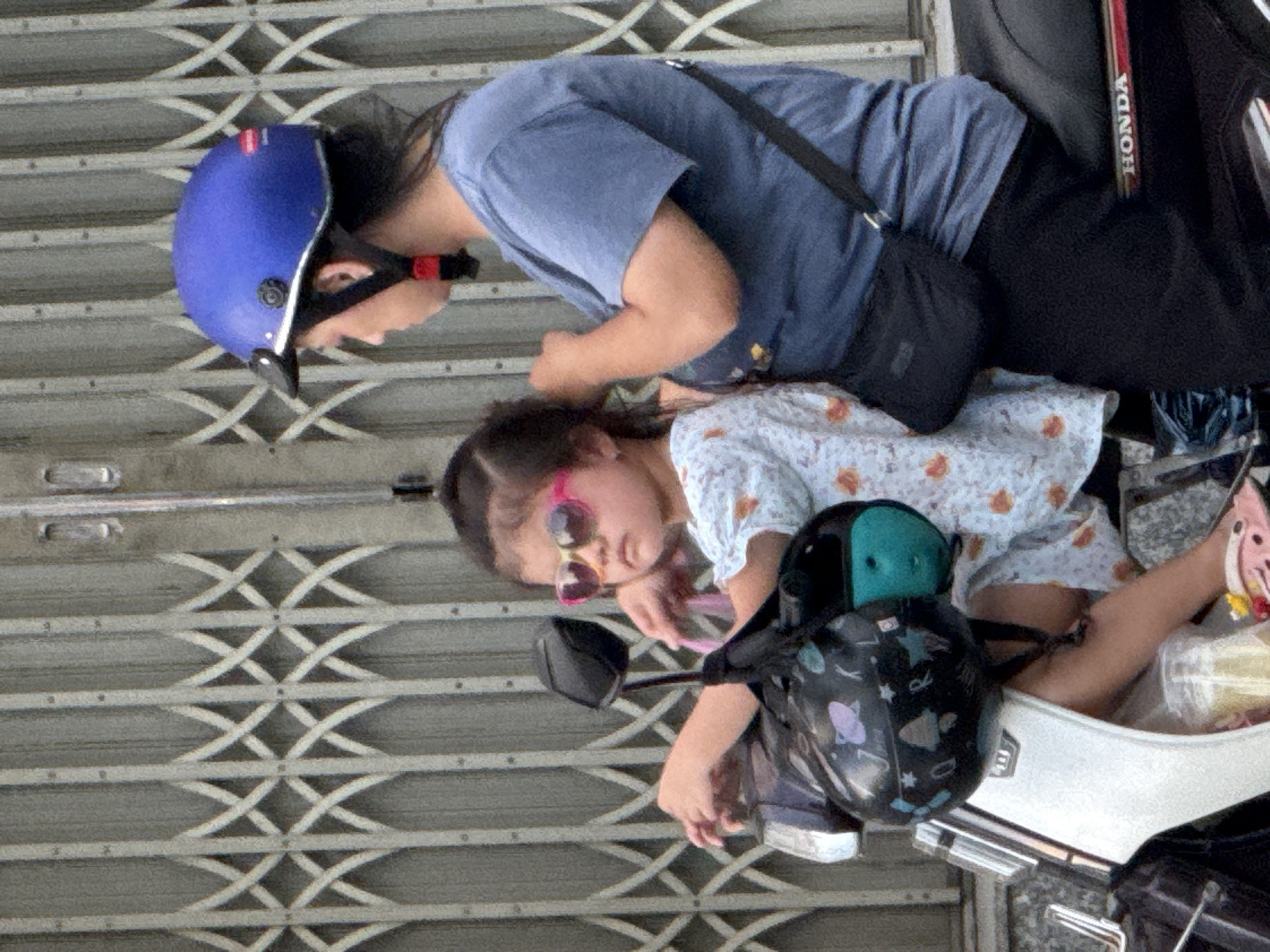 Two individuals, an adult and a child, are seated on a white and black motorcycle in front of a metal grate in Nha Trang, Vietnam. The adult, wearing a blue t-shirt and black pants, has a blue helmet on their head and is adjusting the child's hair. The child, wearing a light blue and white patterned shirt and pink sunglasses, is looking to the left. A black helmet adorned with space-themed designs is placed on the motorcycle in front of the child. A black bag is slung across the adult's body. The environment appears to be an urban setting, possibly a street, with a metal gate providing a textured backdrop.