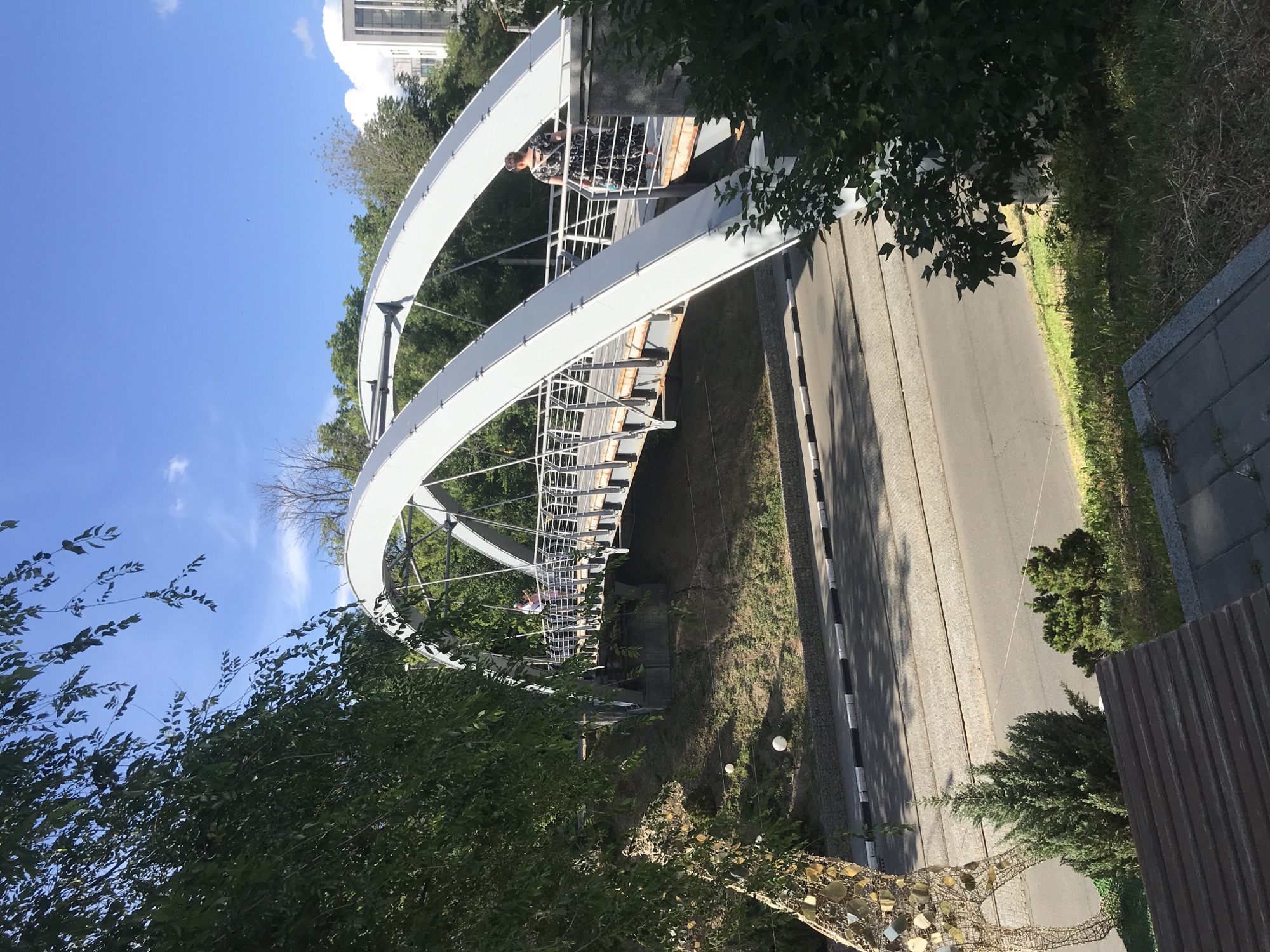 The image shows a pedestrian bridge with two arched supports over a road. A woman in a dark, patterned dress stands on the bridge, facing away from the camera. She is holding a light-colored bag and a blue object. Another person, whose gender is not clear, is visible further down the bridge, wearing a pink shirt and dark pants. The bridge has a metal railing. The surrounding environment includes lush green trees and foliage, a paved road with black and white markings, and a decorative wire sculpture of a giraffe on the left side of the frame. The sky is blue with scattered white clouds, and sunlight casts shadows on the road. In the background, a modern building can be seen. The scene appears to be in an urban park or recreational area.