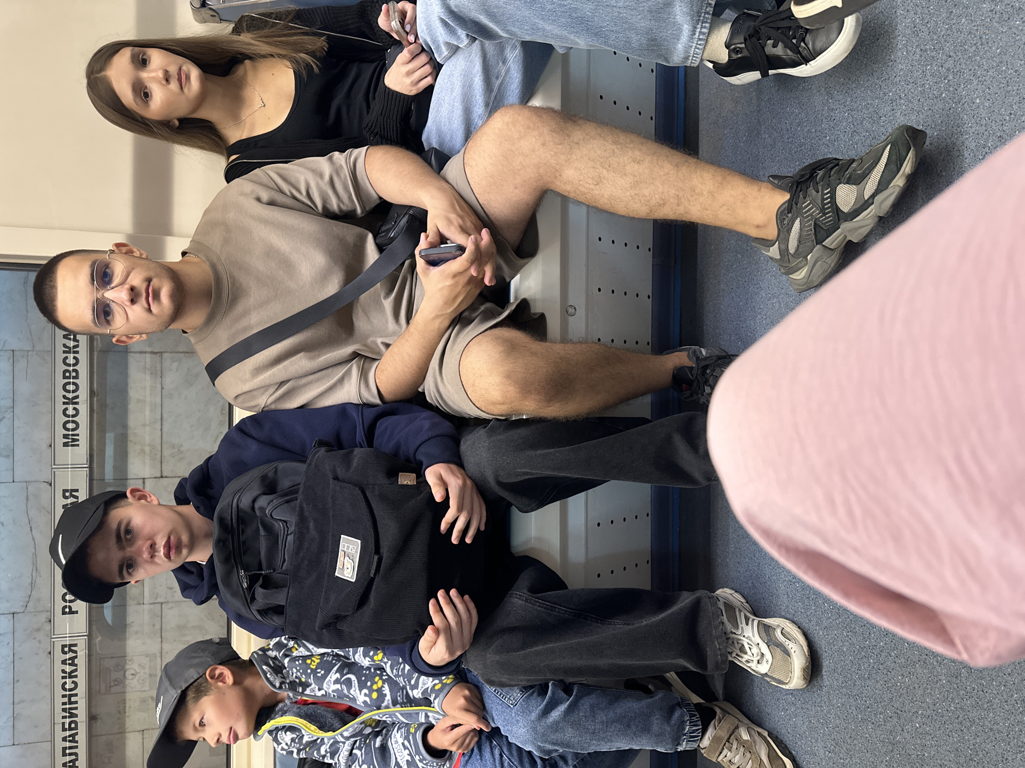 The image shows five young people sitting on a bench in what appears to be a subway car. The bench is metal and has rows of small circular indentations. Above them, white signage with black lettering is visible, including the words "АЛАБИНСКАЯ", "РО", and "МОСКОВСКАЯ", indicating a Moscow Metro station.
From left to right, the first person is a boy wearing a dark baseball cap, a patterned jacket with yellow and white accents, and blue jeans. He is looking to his left.
Next to him sits a teenage boy wearing a black baseball cap, a dark blue hoodie, and dark grey jeans. He is holding a black backpack on his lap with both hands. His gaze is directed forward and slightly to his left.
To his right is a young man with short dark hair and glasses, wearing a light brown crewneck sweatshirt and matching shorts. A dark strap is slung across his chest. He is holding a smartphone in his right hand.
Beside him is a young woman with long brown hair, wearing a black tank top and a dark cardigan. She has a delicate necklace with a small pendant. She is holding a smartphone in her right hand.
The far right of the image is partially obscured by a pink garment in the foreground. A person's legs and feet are visible wearing light blue jeans and black and white sneakers.
The overall lighting suggests an indoor environment, consistent with a subway car. The people are casually dressed and appear to be traveling.