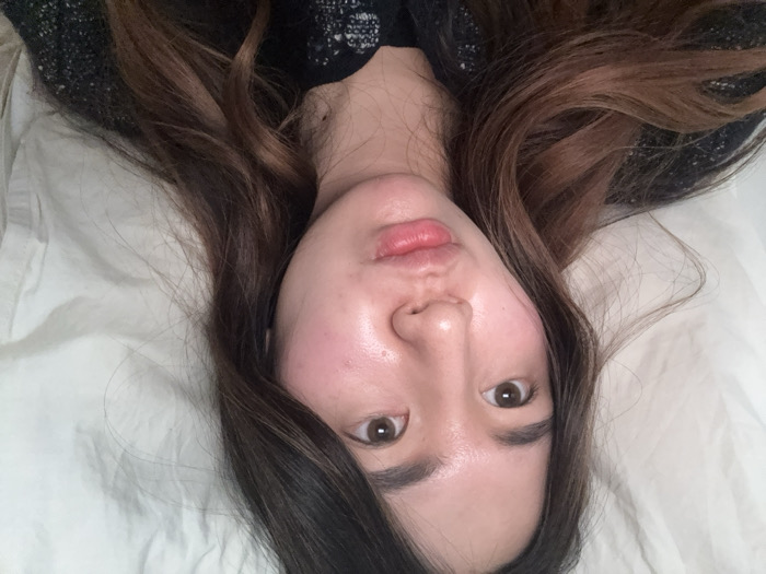 A young East Asian female is depicted in a close-up, chest-up shot. Her long, wavy dark brown hair, featuring lighter highlights, frames her face. Her medium-dark brown eyes are open and directed towards the viewer. Her lips are pursed in a slight pout. Her complexion is fair with a natural sheen and a subtle pinkish flush. She is wearing a dark, textured garment. The background consists of uniformly light-colored, crumpled fabric, consistent with bedding or a draped sheet. No identifiable landmarks or specific environmental cues indicating a location in Paris, France, or any other city are present.