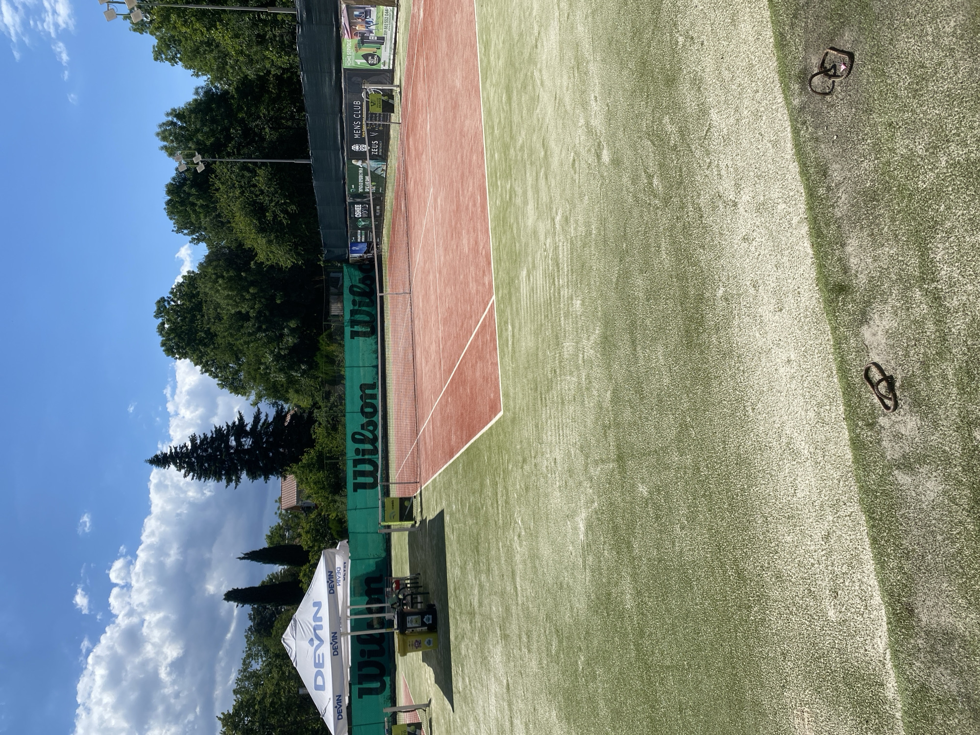 This media file depicts a tennis court on a sunny day. The court has a red clay surface on one side and a green artificial turf on the other, separated by a net. A green banner with "Wilson" written on it runs along the fence. There are advertising banners on the fence as well, one of which reads "MEN'S CLUB" and another with "DEVIN". Two white tents are pitched near the court, also bearing the "DEVIN" logo. In the background, lush green trees and a clear blue sky with fluffy white clouds suggest a pleasant outdoor setting. There are also some metal rings visible on the green surface of the court. The image appears to be taken from a slightly elevated perspective, looking down onto the court. There is no visible activity or people on the court itself, suggesting it is either before or after a game, or perhaps being used for practice. The lighting suggests it is daytime, likely late morning or early afternoon.