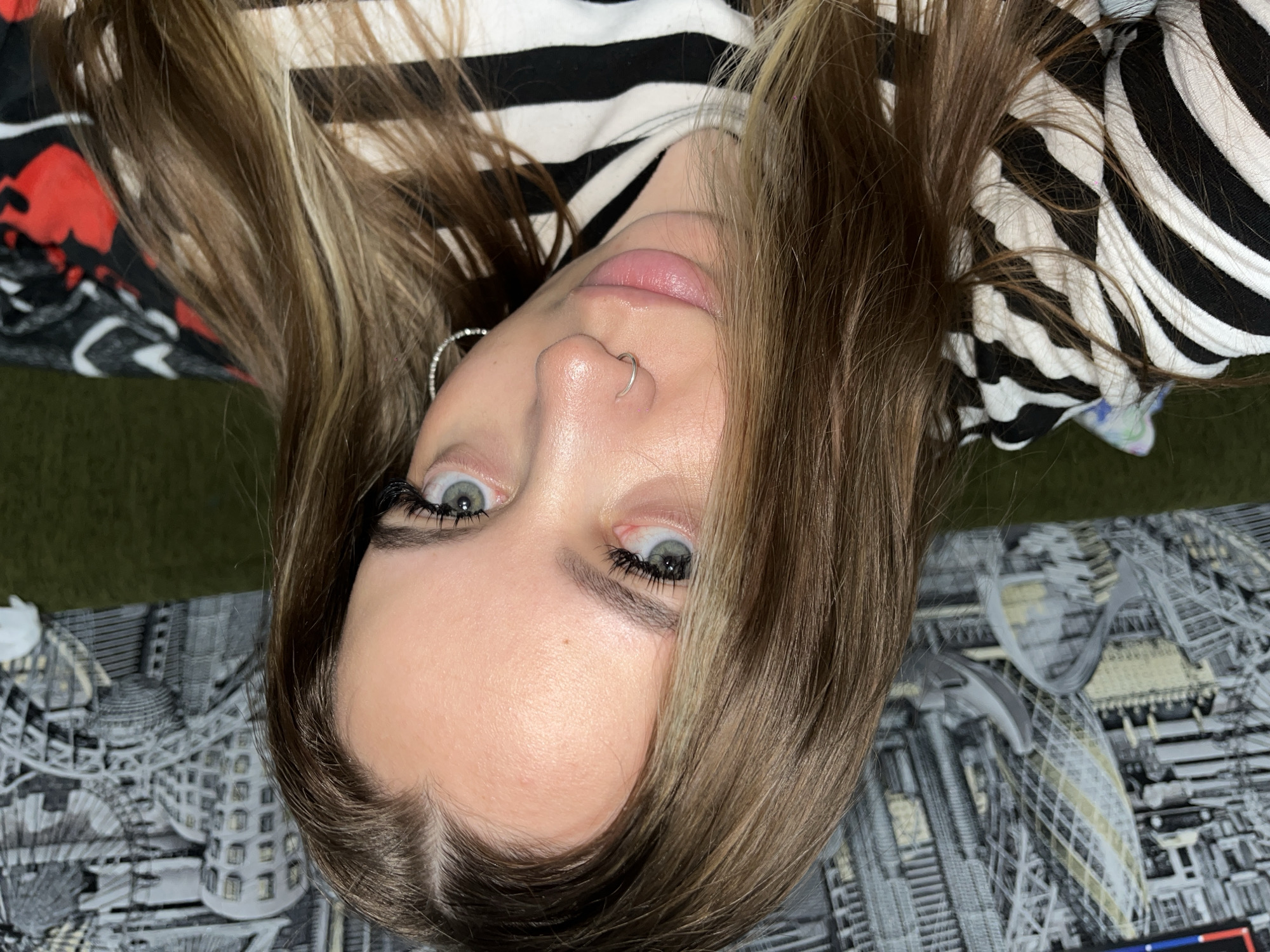 A woman with light brown hair and green eyes is pictured looking directly at the camera. She has a nose ring and is wearing eyelash extensions. She is wearing a black and white striped shirt. The background includes a patterned blanket with a cityscape design and a dark green surface. There is no indication of location (Novopokrovskaya, Russia).