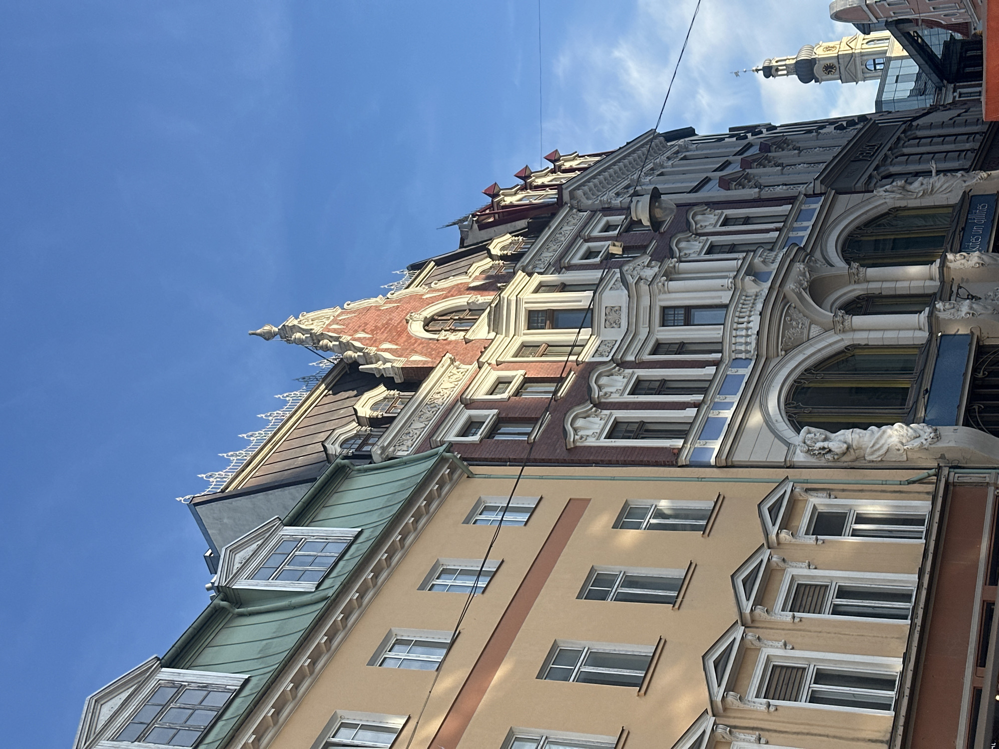 The image shows a street view of buildings in Riga, Latvia, known for its Art Nouveau architecture. The buildings display ornate facades with varying colors, including beige, red brick, and green roofs. Decorative elements like sculptures, arches, and intricate window designs are visible. The sky is clear and blue. A church tower can be seen between buildings.