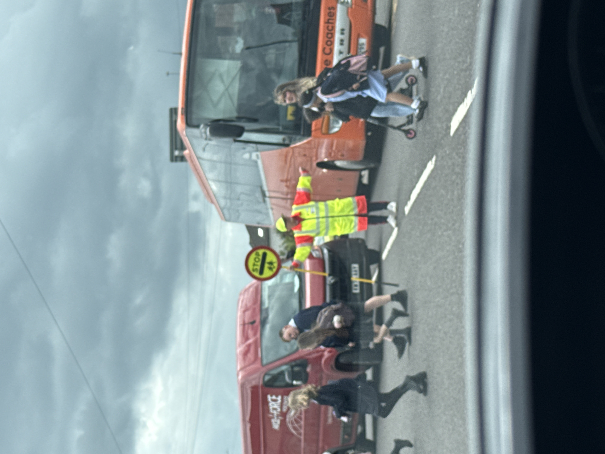 The image shows a street scene in what appears to be Great Yarmouth, United Kingdom. A person in a high-visibility jacket and hat is holding a "Stop" sign to allow school children to cross the street. Several children in school uniforms are crossing the street. There is a red van and an orange bus or coach stopped on the road. The sky is overcast. The image is taken from inside a vehicle. A girl is on a scooter crossing in front of the bus.