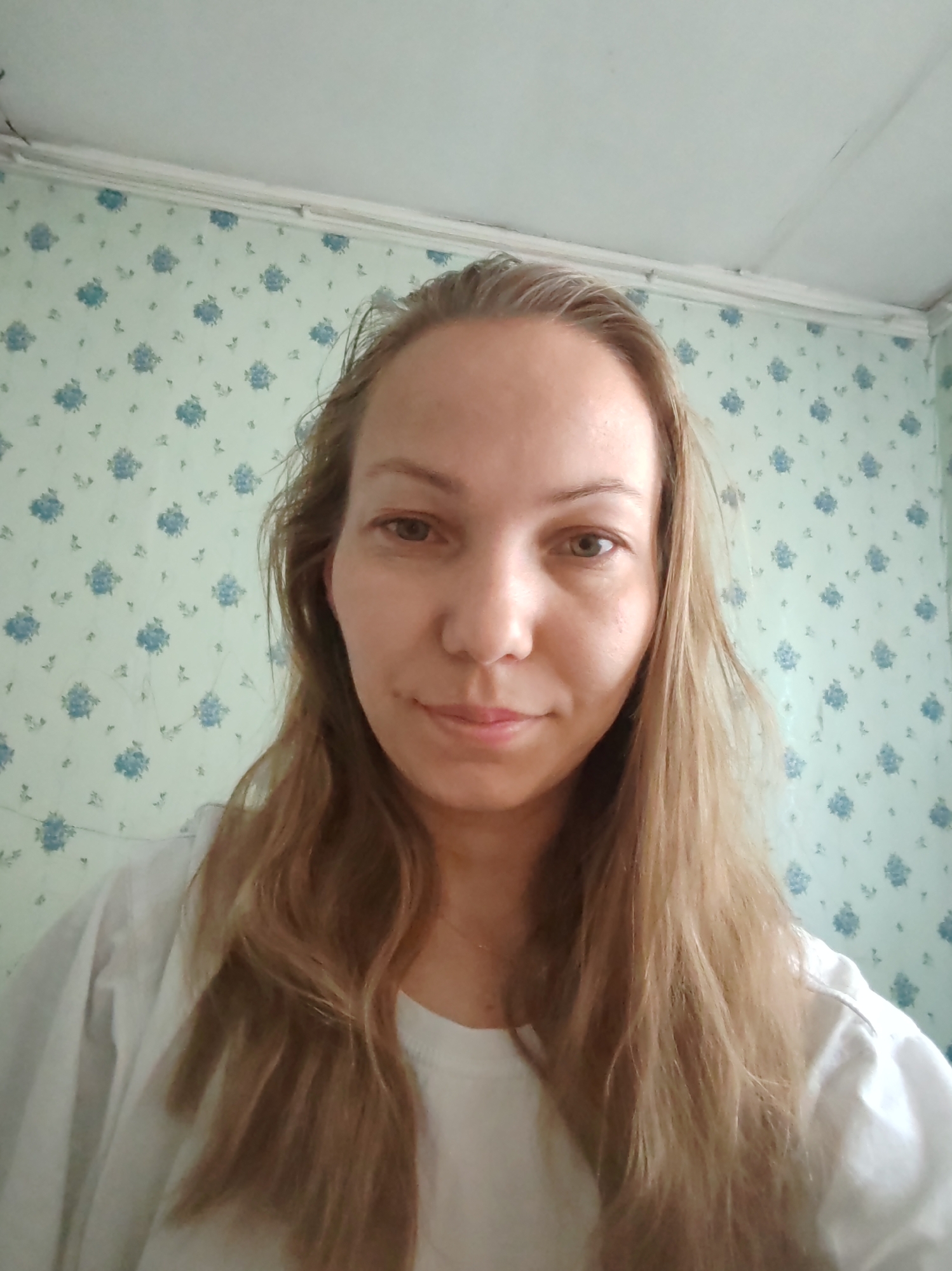 A medium shot shows a Caucasian woman with long, light brown hair and fair skin. She is looking directly at the camera with a neutral expression. She is wearing a white t-shirt. The background consists of a light blue floral wallpaper and a white ceiling. The image appears to be a selfie, taken indoors in what seems to be a residential setting. The specific location of Чухлэм, Russia, is not visually discernible from the image itself.