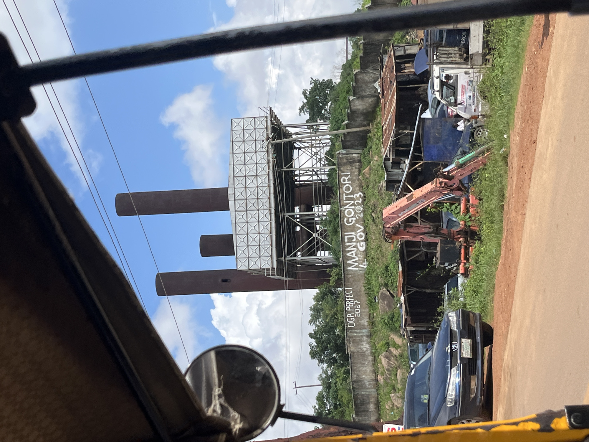 The image, taken from inside a yellow motorized rickshaw, shows a view of an industrial site in Jos, Nigeria. In the background, three tall, dark smokestacks rise against a bright blue sky with scattered white clouds. A large, metallic water tank structure with a criss-cross pattern sits between the smokestacks. Below this industrial structure, a concrete wall is visible, with graffiti spray-painted on it. The graffiti reads "OGA PERFECT 2027" and "MANJI GONZORI GOV 2023". The foreground of the scene depicts a collection of various vehicles and structures. A dark blue sedan is parked on a dirt road. Behind it, an orange excavator arm is visible, along with several makeshift shacks with corrugated metal roofs. A white truck is also present, with two individuals seated inside. The surrounding area is covered in green vegetation and rocky terrain.