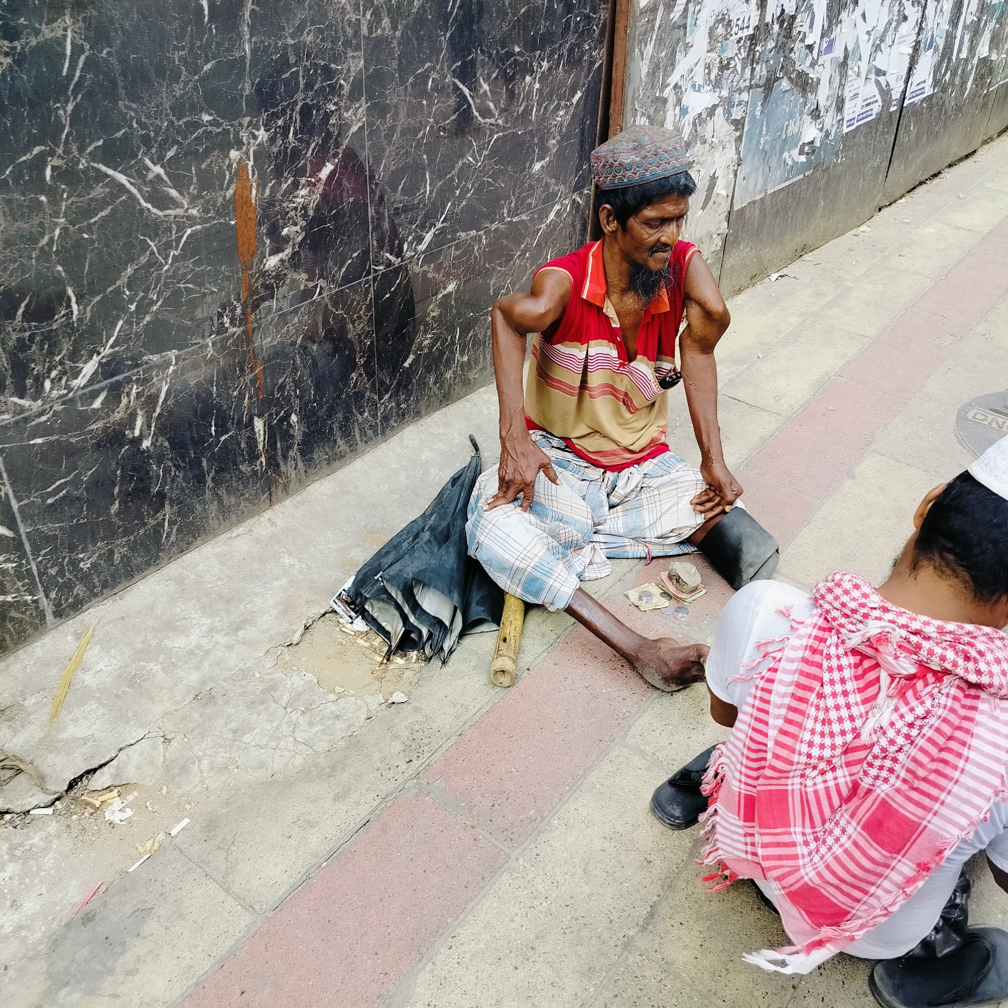 This image captures a street scene in Dhaka, Bangladesh, featuring two men sitting on a concrete sidewalk during the daytime.
The prominent subject, positioned on the left, is a man with dark skin, a dark beard, and a mustache. He wears a patterned skullcap (kufi), a red and beige striped polo shirt, and a light blue and white checkered lungi. He exhibits notable physical disabilities, with visibly shortened and bent arms, and his legs also appear affected; his right foot rests on the ground while his left leg is bent near a dark, possibly prosthetic, support. A small pipe or cigar is held near his mouth. In front of him, various coins, some stacked on a small stone, are spread on the ground, suggesting he is collecting alms. A collapsed black umbrella and a bamboo stick lie beside him. His gaze is directed downwards or to the side, giving him a neutral or somber expression.
The second man, seen partially from the back on the right, wears a white shirt, a white skullcap, and a distinctive red and white checkered keffiyeh-like scarf draped over his shoulders. He is also seated on the ground, facing the first man.
The background on the left features a dark, reflective wall with prominent white marble-like veining and hints of brown coloration. To the right, behind the two men, is a lighter-colored wall heavily plastered with numerous torn and faded posters and flyers, some with indistinct text, including a partially visible "544". The ground is primarily light grey concrete, showing cracks and scattered debris like cigarette butts. A distinct strip of reddish-brown pavers is embedded in the concrete pathway. A faint "DNA" inscription is visible on a circular utility cover on the ground. The bright, even lighting indicates it is daytime, and the scene depicts an everyday urban setting.