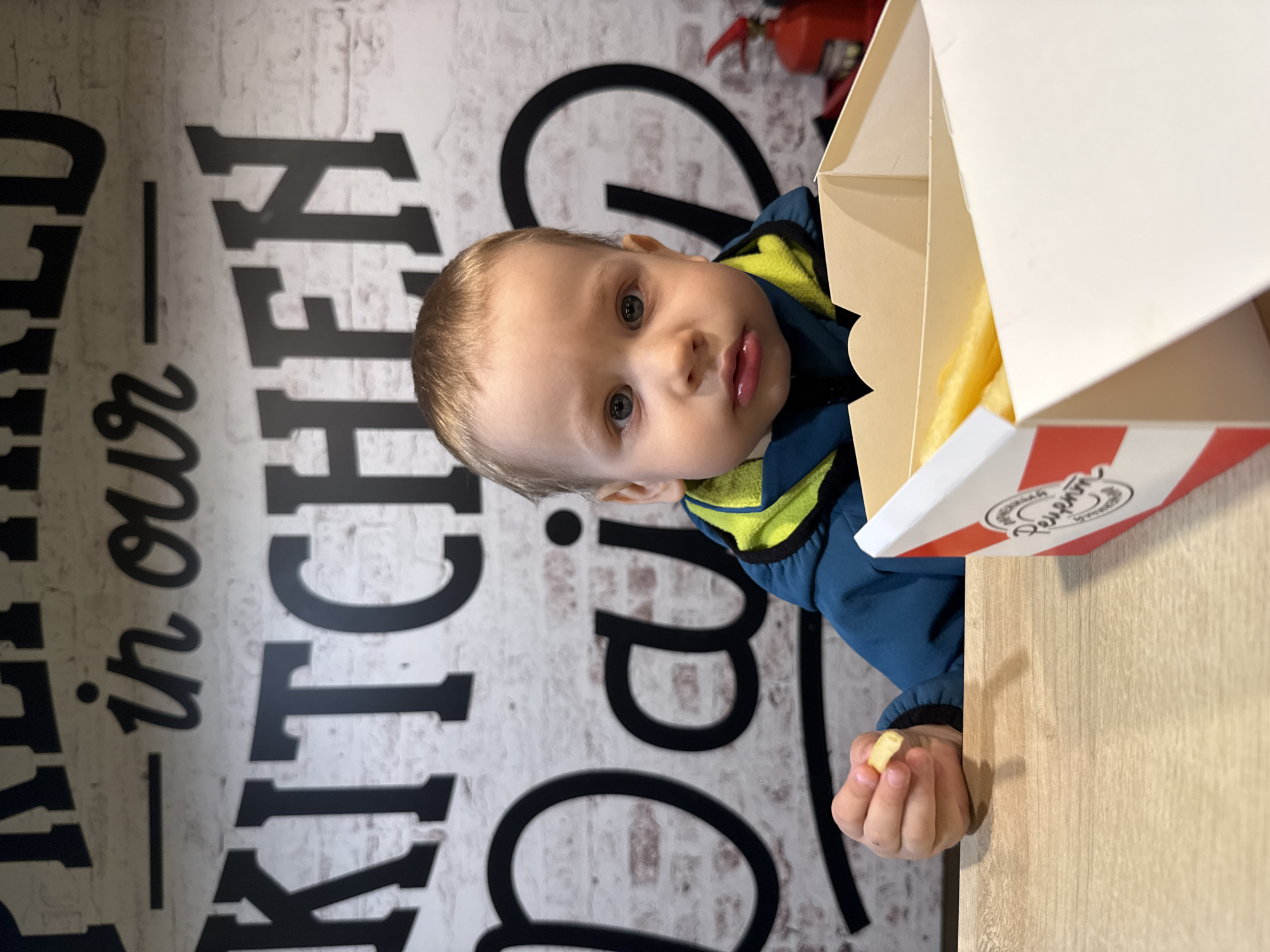 A young child sits at a wooden table next to a box of french fries. The child is wearing a blue jacket with a bright yellow lining. In the background is a wall with the words "Held in our Kitchen" printed in black lettering. A fire extinguisher is partially visible in the upper right corner of the image. The side of the french fry box has a red and white design, with Russian text. It is impossible to determine if this is Rusanovo, Russia, based on the image alone.