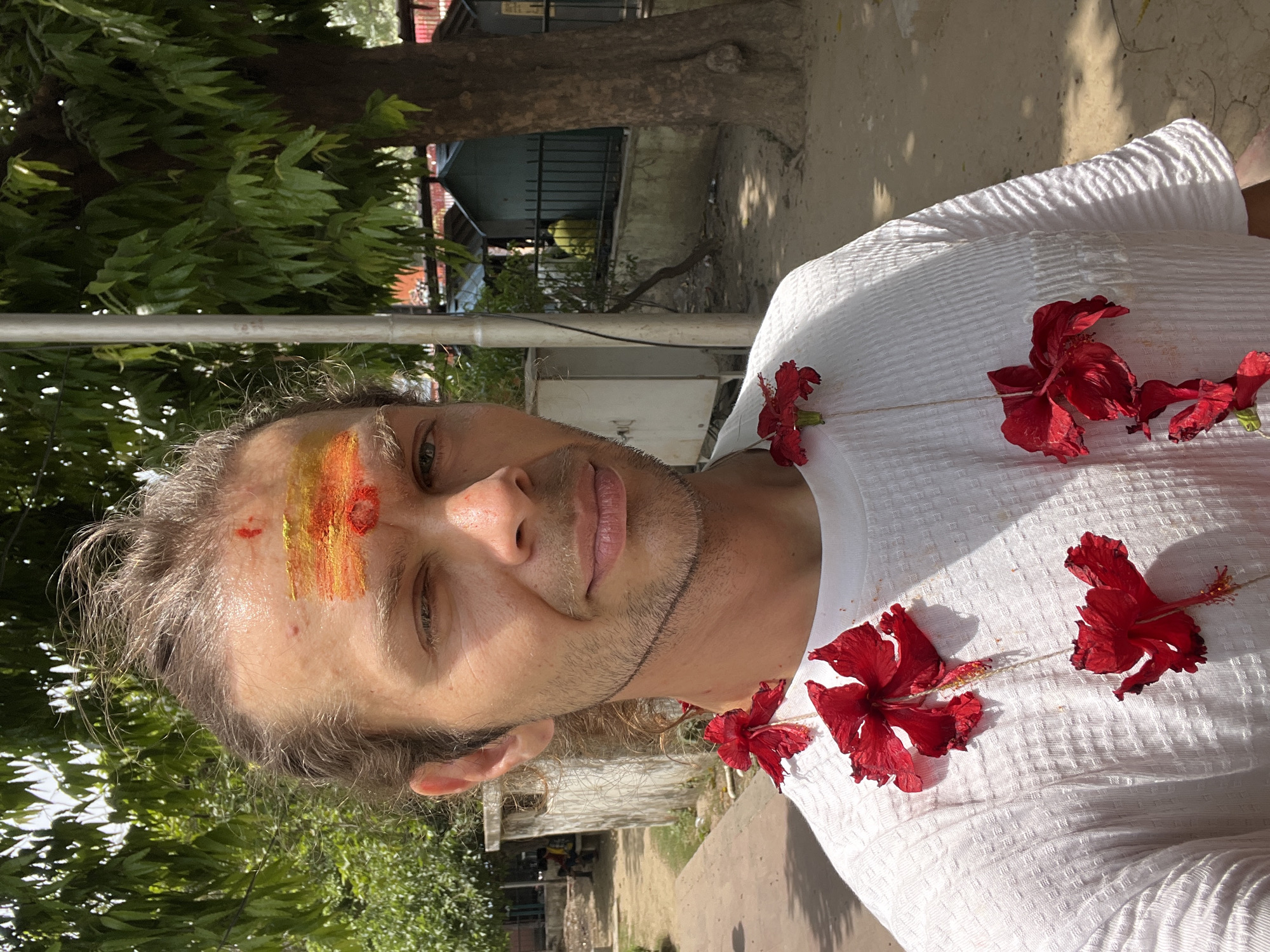 A fair-skinned man with light brown hair and stubble is the primary subject of this close-up, head-and-shoulders shot. He wears a white, textured T-shirt adorned with several large, red hibiscus flowers. A streak of yellow and orange paint is smeared across his forehead. The background appears to be an outdoor urban setting. The location is Varanasi, India. Visible elements include foliage, trees, buildings, and a metal pole. The image is rotated counter-clockwise.