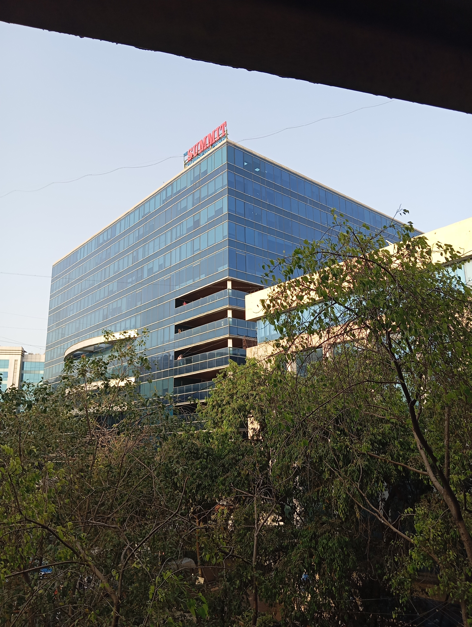 The image depicts a tall, modern office building with a glass facade. The building is located in Mumbai, India, as indicated by the sign on top that reads "THE SUMMIT BUSINESS BAY." The building has multiple floors, and some floors have balconies. Trees partially obscure the lower part of the building. The sky is light blue. The photograph is taken from a slightly elevated position, possibly from a window or balcony of another building.
