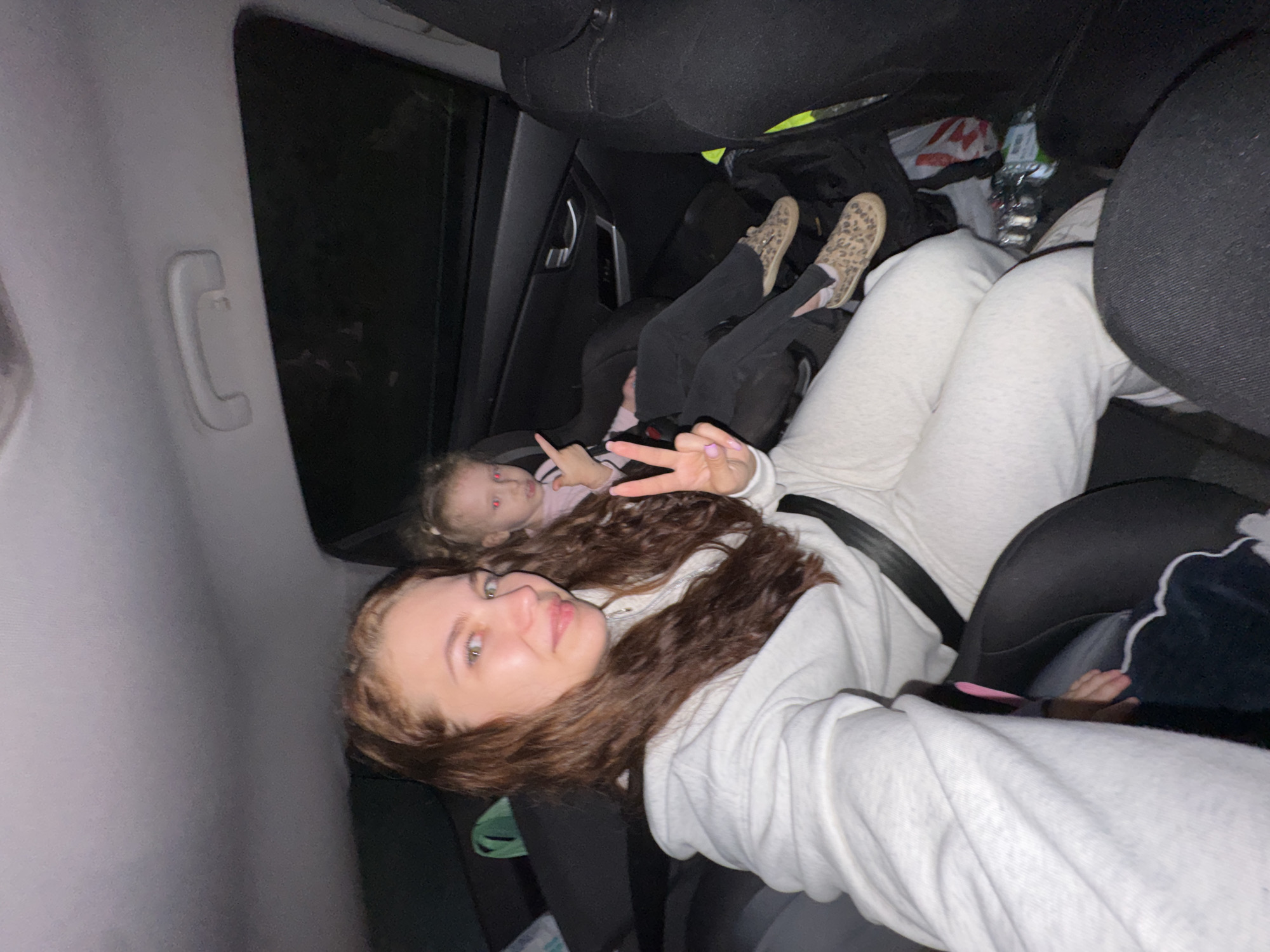 The image shows the interior of a car, likely taken from the passenger seat looking towards the back. Three individuals are visible. A young woman with brown hair is in the foreground, wearing a white sweatshirt and a seatbelt. She is holding up two fingers in a peace sign. Behind her, a child in a car seat is visible, with a small hand and bare foot visible. Further back, another child can be seen sitting, wearing black leggings and animal print shoes. The car is dark, with a water bottle and plastic shopping bag visible on the back seat. The environment appears to be enclosed, suggesting the inside of a vehicle, possibly at night or in a dimly lit area. There is no information to suggest the location is in Chaikovsky, Russia.