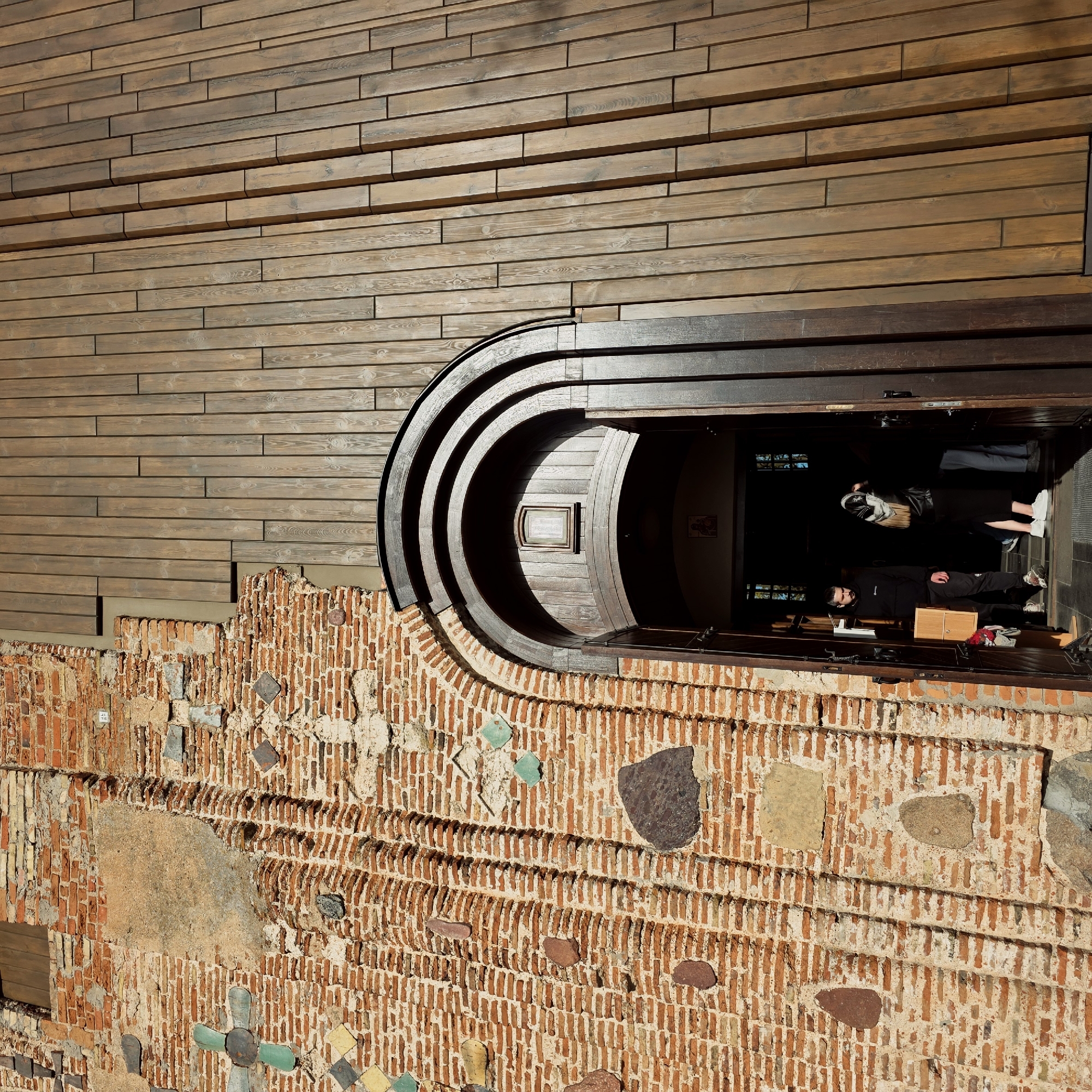 The image displays a unique building facade in Hrodna, Belarus, highlighting a compelling blend of historical and contemporary architecture under clear, bright daylight. The upper part of the structure is clad in a modern wooden facade, composed of horizontal planks of varying lengths and depths, which creates a textured, stepped appearance.
Below this wooden cladding, an ancient-looking brick wall forms the base of the building. This wall features rustic, reddish-brown bricks laid in various patterns, interspersed with irregularly shaped stones and decorative elements such as small square tiles, some green and others patterned, contributing to its intricate and aged character.
A distinctive, deep, semicircular archway serves as an entrance, seamlessly transitioning between the wooden facade and the brickwork. This archway is constructed from dark wood, with multiple concentric rings creating a visually striking, layered tunnel effect. Inside the dimly lit entrance, two individuals are visible. A man with a beard, dressed in dark clothing, stands behind a counter or ledge, facing outwards. Another person, partially visible with light pants and white sneakers, stands further back within the entrance. A small, framed object, likely a sign or menu, is affixed to the inner curve of the wooden archway. The strong sunlight casts prominent shadows, emphasizing the architectural details and the depth of the entrance, showcasing a harmonious fusion of traditional and modern design.