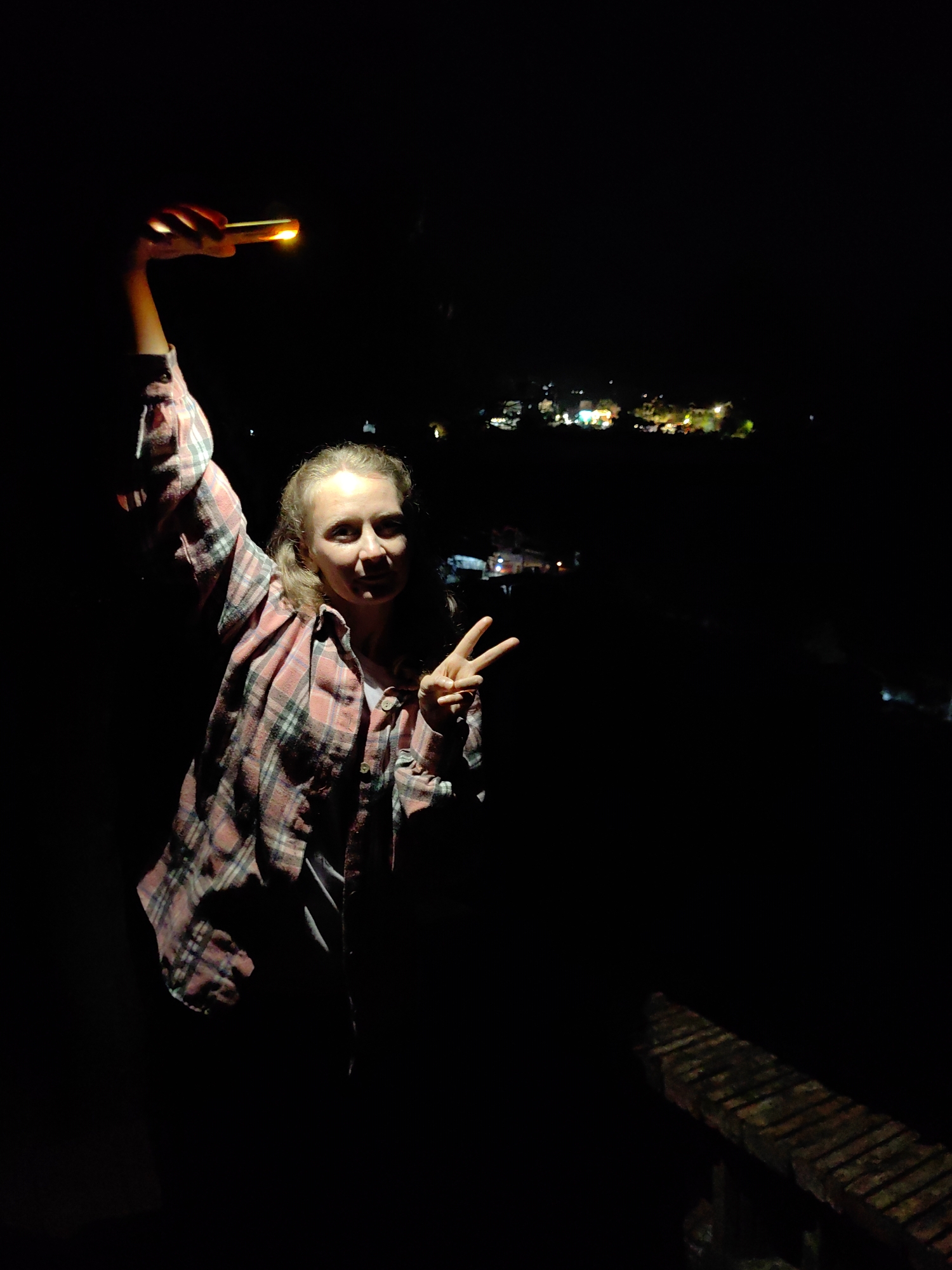 At night in Xã Đàm Thủy, Vietnam, a woman stands on a weathered wooden surface, possibly a bridge or platform. She is holding a lit cell phone aloft in her right hand and making a "peace" sign with her left hand. She is wearing a long-sleeved, pink and grey plaid shirt over a white shirt. In the dark background, there are distant lights from what appears to be a settlement or town.