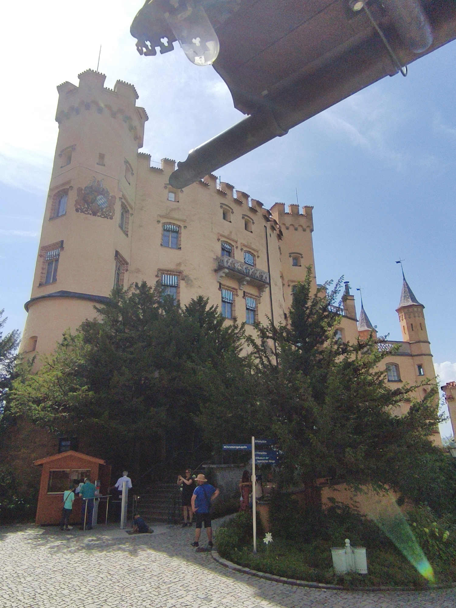 A low-angle, full shot captures the imposing façade of Hohenschwangau Castle in Schwangau, Germany, under a bright, clear sky. The castle, a towering structure of pale yellow, dominates the upper portion of the frame, its crenellated turrets and numerous windows hinting at its historical significance. A large, dark, metallic object, possibly part of an overhead structure or vehicle, intrudes into the top of the image, casting a shadow that partially obscures the upper parts of the castle.
In the foreground, a cobblestone path leads towards the castle. A small, reddish-brown wooden ticket booth is positioned to the left, with several people gathered around it. A person in a blue shirt and dark shorts stands facing away from the camera, looking towards the castle. Another person in a white shirt and dark pants stands by a turnstile, appearing to be an attendant. Two people are visible near the ticket booth, one in a turquoise shirt and dark pants, and another in a green shirt and dark pants, seemingly interacting with the booth.
To the right of the path, a lush, dark green tree with dense foliage partially obscures the base of the castle. Behind the trees and to the right, a pathway leads upwards towards the castle, with a signpost indicating directions. Several figures are seen on this elevated path, including a woman in a black dress taking a photo with her phone, and another person in a dark top and light pants.
The environment is characterized by a sunny day with a clear blue sky, interrupted only by the looming presence of the metallic object. The overall impression is one of a historic site bustling with tourists on a pleasant day.