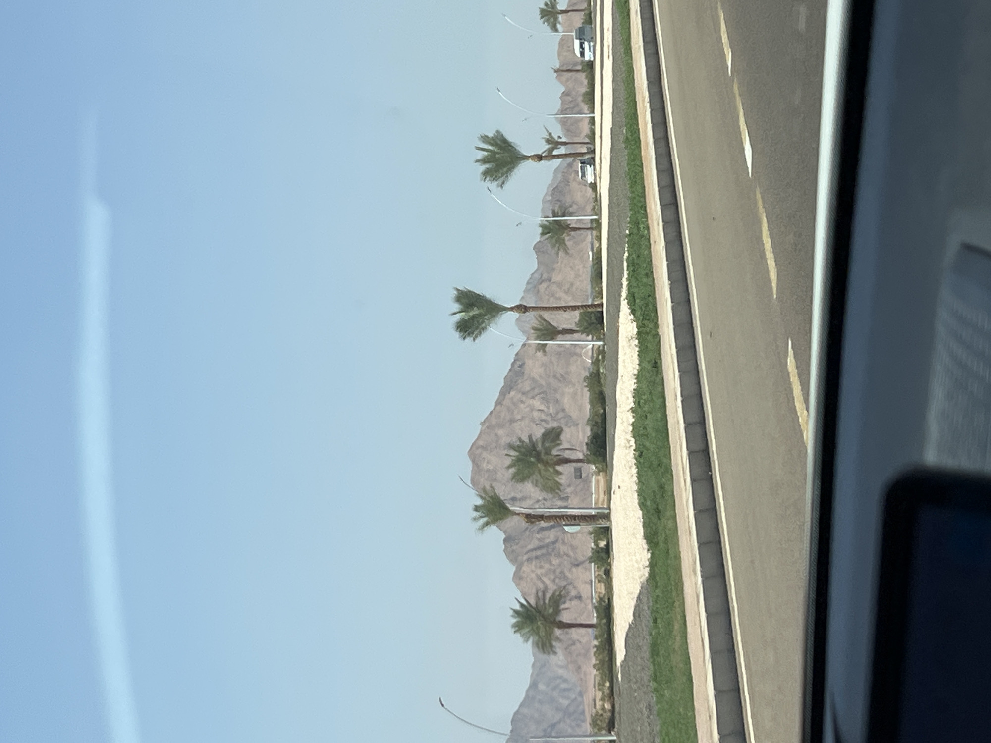 The image shows a road in Sharm El Sheikh, Egypt, with palm trees and mountains in the background. The road is paved with asphalt and has yellow lane markings. There are several palm trees lining the road, with lush green grass and sand in between. The mountains in the background are rocky and arid, typical of the desert landscape of the region. The sky is clear and blue, indicating a sunny day. There are no people visible in the image, but there are a few vehicles parked along the road. The scene suggests a well-maintained area with landscaping, possibly a residential or tourist area.