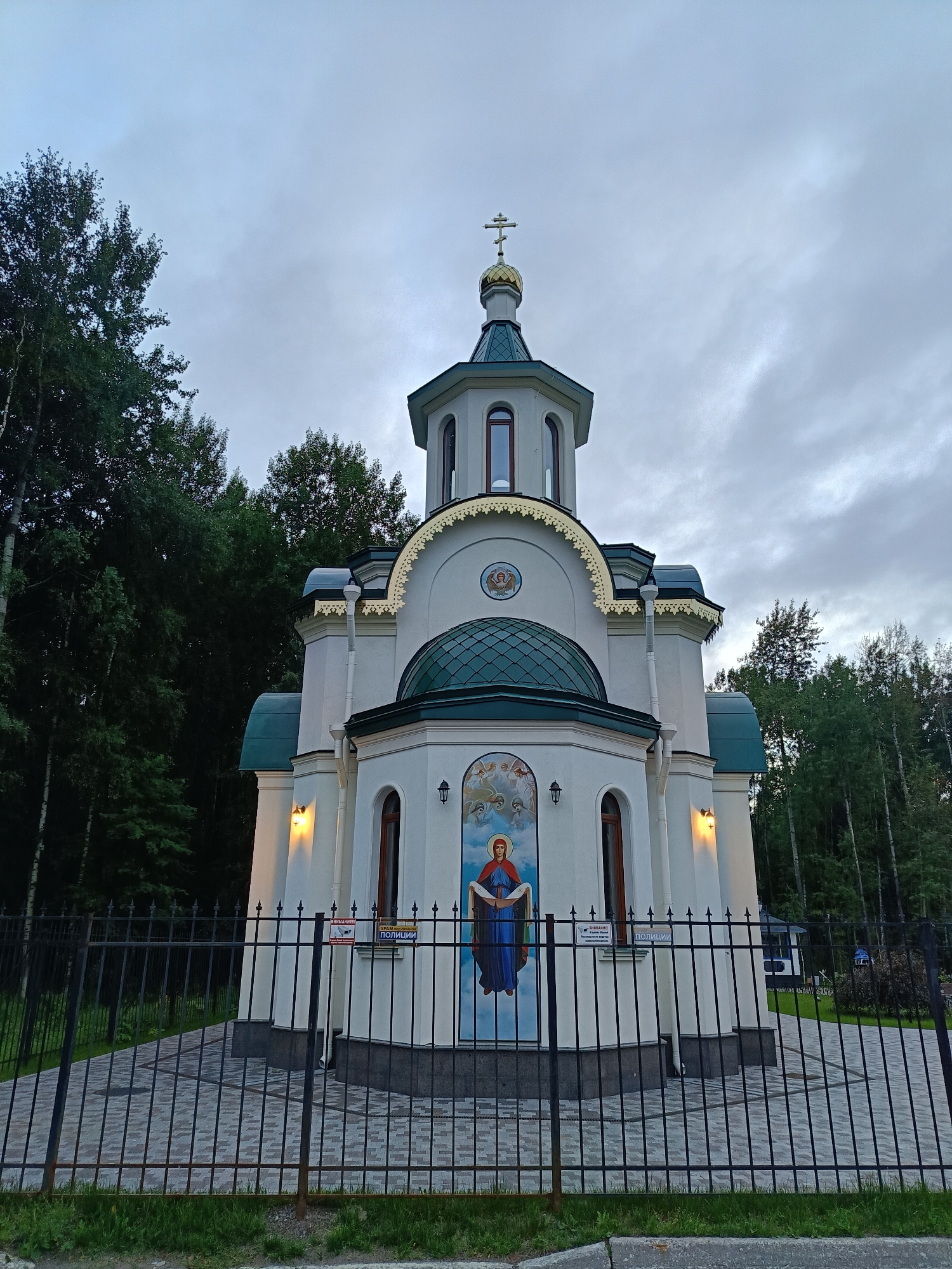The image depicts an Orthodox chapel located in Saint Petersburg, Russia, nestled amongst dense greenery. The chapel features a white facade adorned with a large fresco of the Virgin Mary on the front, flanked by angels. It has a distinctive green dome topped with a golden cross, and smaller green roof sections. Decorative golden detailing accents the roofline. A black metal fence encloses the chapel and its immediate surroundings, which are paved with cobblestones. The sky above is overcast and grey, suggesting a cloudy day. The area around the chapel is heavily wooded, with tall trees forming a natural backdrop. The chapel is illuminated by exterior lights, some of which are visibly lit. Signs are present on the chapel wall, one indicating "POLITSIYA" (Police).