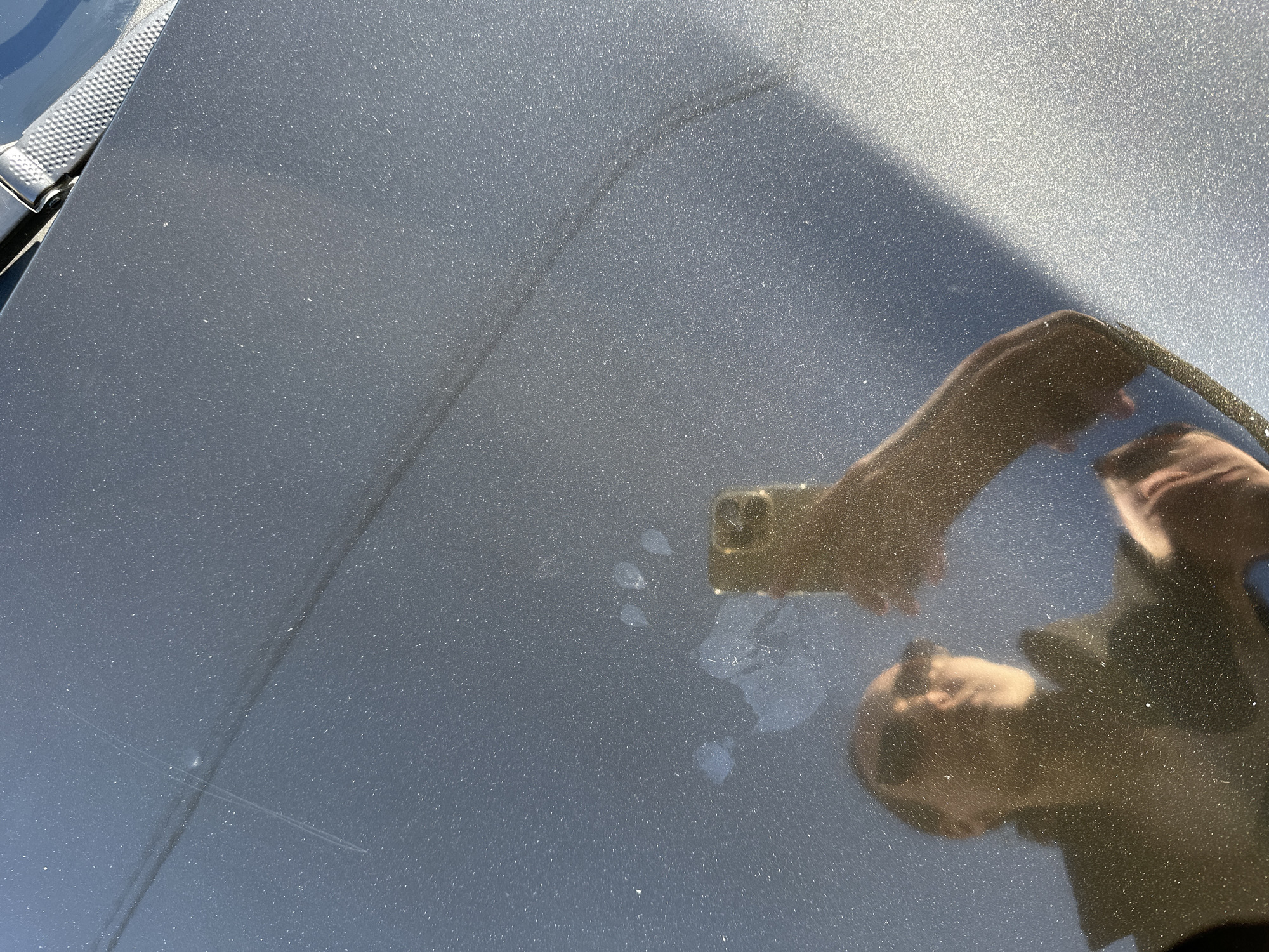 The image shows a close-up of a metallic blue car hood. In the reflection, a person is visible holding a smartphone, presumably taking the picture. There are some water spots or dried residue on the hood. The overall scene is brightly lit, suggesting an outdoor location. There is no information in the image that indicates a location in Подшивалово, Russia.