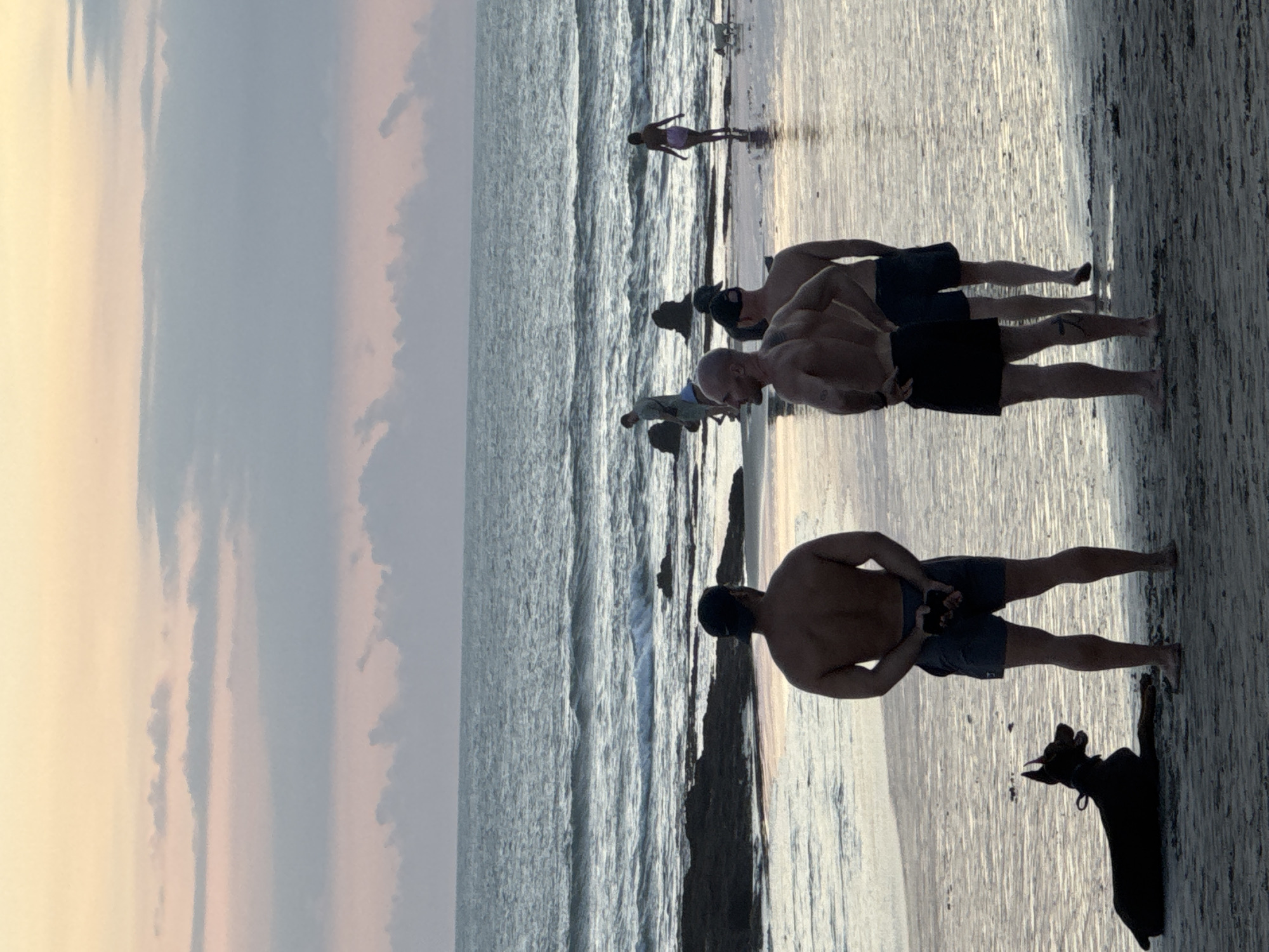 The image captures a beach scene in North Kuta, Indonesia, during either sunrise or sunset, evidenced by the warm tones of the sky on the left side. The ocean, with visible waves, occupies the central portion of the image. On the right side of the image, a group of people are standing on the wet sand. The foreground features a man wearing dark shorts and a cap, standing with his back to the camera. A dog is also present near him, casting a long shadow on the wet sand. Further back, there are two more men, shirtless and wearing shorts, with one wearing a hat. Further in the distance, there are two additional individuals, with one person walking by the waters edge.