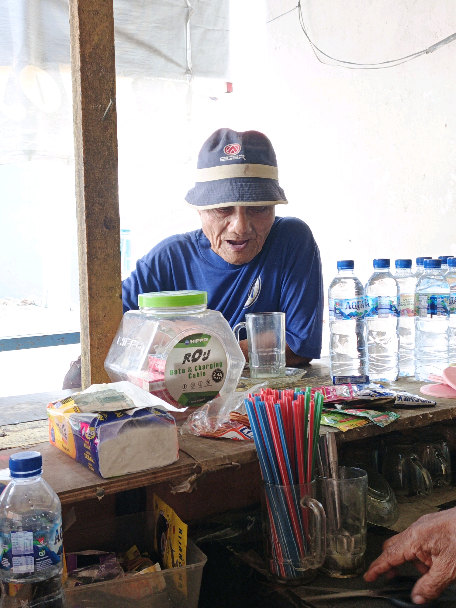 An elderly man wearing a dark blue bucket hat with a beige band and a dark blue t-shirt looks down at a wooden counter. He is in what appears to be an outdoor market stall, likely in Ketegan, Indonesia, indicated by the text on a water bottle. The stall has a rough wooden post on the left, a white tarp or ceiling above, and a white textured wall behind.
On the counter in front of the man are several items. To his left is a large, clear plastic jar with a green lid, containing what looks like packaged goods. In front of the jar is a package of white tissues. To the right of the jar are several bottles of AquA water, lined up, some partially obscured by others. A clear glass mug is placed to the left of the water bottles. In front of the glass mug and water bottles is a collection of brightly colored drinking straws, red, blue, and green, held upright in a clear glass. To the right of the straws, another clear glass is visible.
The man's mouth is slightly open as if speaking or about to speak. His hands are not visible in the frame. The overall lighting suggests daylight. The environment appears functional and somewhat cluttered, typical of a small retail stall. A portion of another person's hand is visible in the lower right corner of the image, pointing towards the drinking straws.