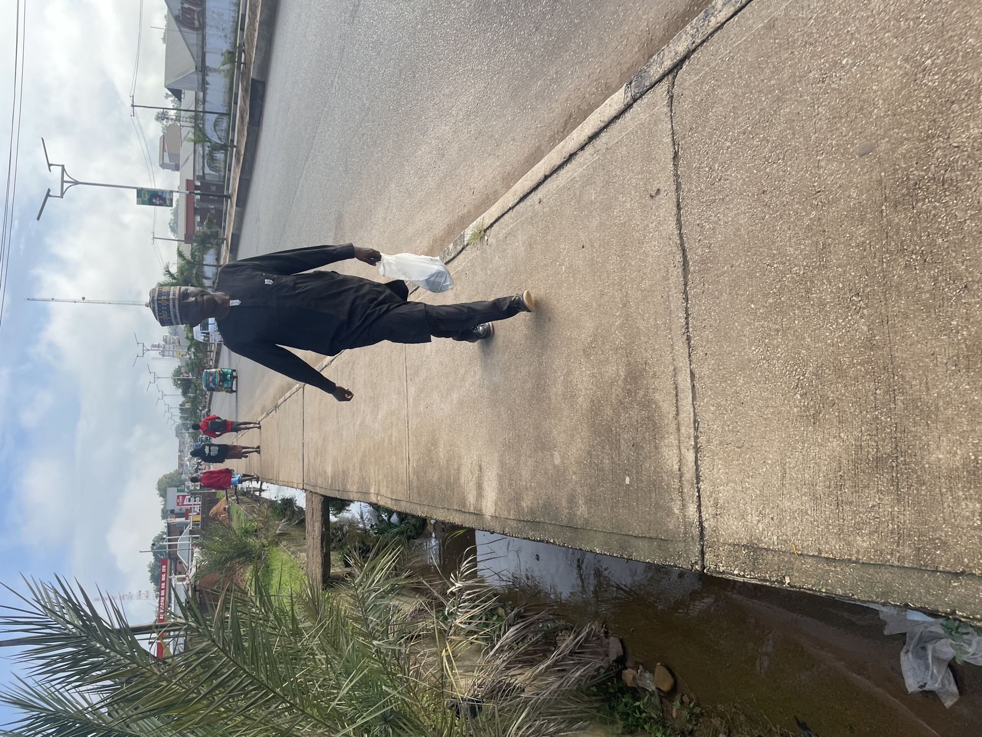 A man wearing a black traditional outfit and a patterned cap walks on a concrete sidewalk next to a drainage ditch. He carries a white plastic bag in his right hand. In the background, several other people, including two men and a woman, walk further down the sidewalk. A three-wheeled vehicle, often called an "okada" or "keke napep" in Nigeria, is visible on the road. The scene appears to be in an urban setting, with buildings and streetlights lining the road. The image captures a moment on a street in Jos, Nigeria, characterized by its typical roadside infrastructure and pedestrian activity.