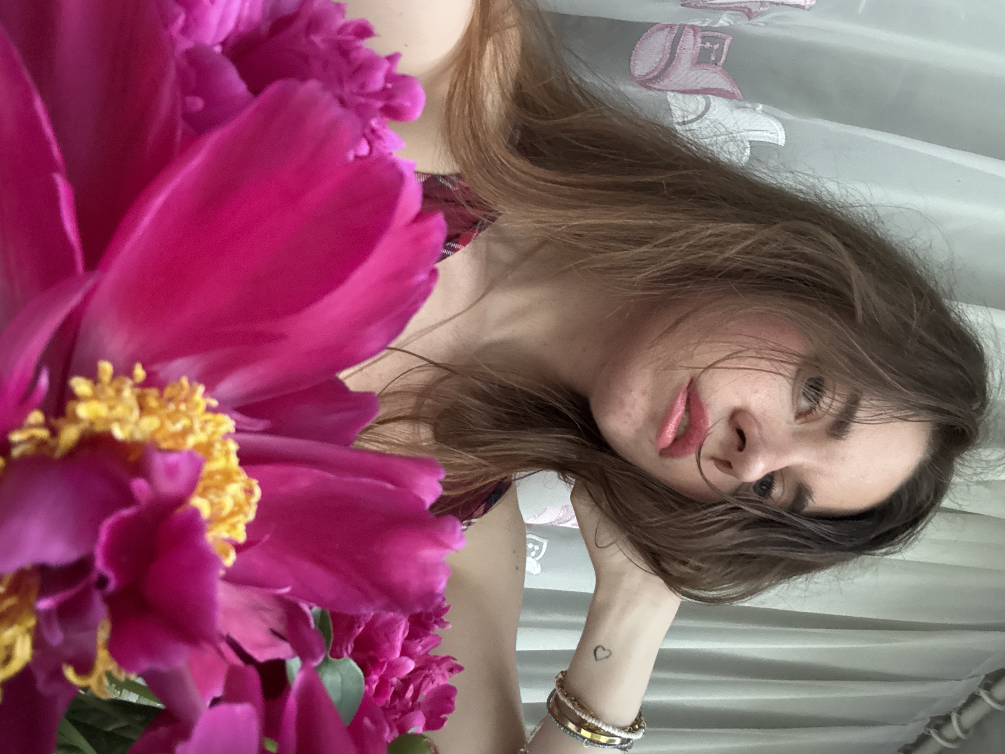 A close-up, medium shot shows a young woman posing for a selfie with a bouquet of pink peonies. Her right arm is bent with her hand resting on the back of her head. She is wearing a plaid top and several bracelets on her right wrist. A small heart tattoo is visible on her left wrist. The background consists of a sheer, white curtain with a pink floral pattern. The woman's hair is brown and falls around her shoulders. The peonies are in the foreground, dominating the lower portion of the frame.