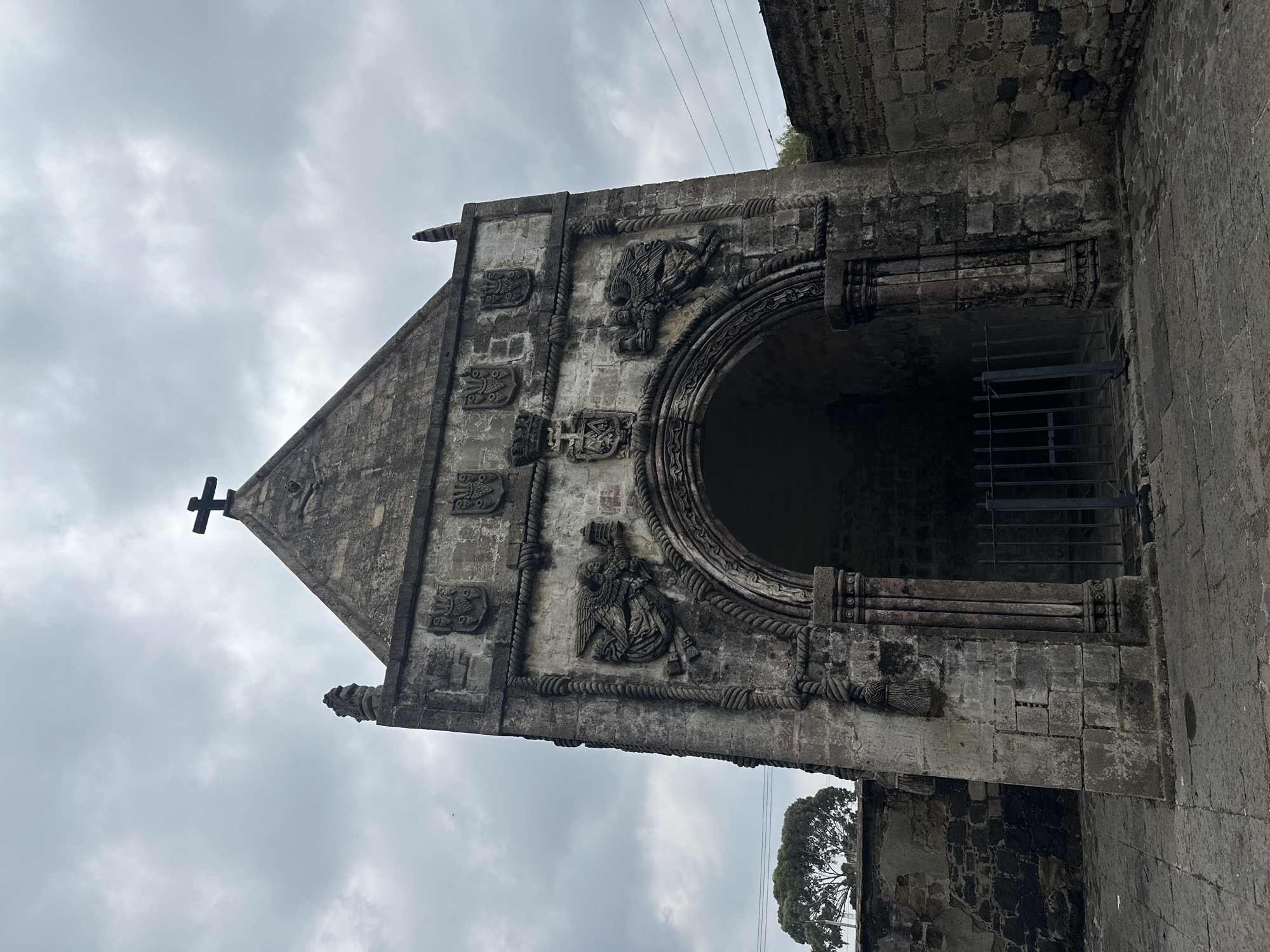 This image shows the exterior of a stone structure, likely a chapel or shrine, with a prominent arched entrance. The structure is made of weathered stone blocks and features intricate carvings. Above the arch, there are depictions of angels holding candelabras and shields with coats of arms. A cross is visible at the apex of the building's triangular gable. The area in front of the entrance is paved with stone, and a dark metal gate is positioned within the archway. To the left of the structure, a stone wall is visible, and further in the background, a single tree stands against a cloudy sky. The overall impression is of an old, historic religious site. This structure is located in Huejotzingo, Mexico, and is part of the Franciscan monastery complex.