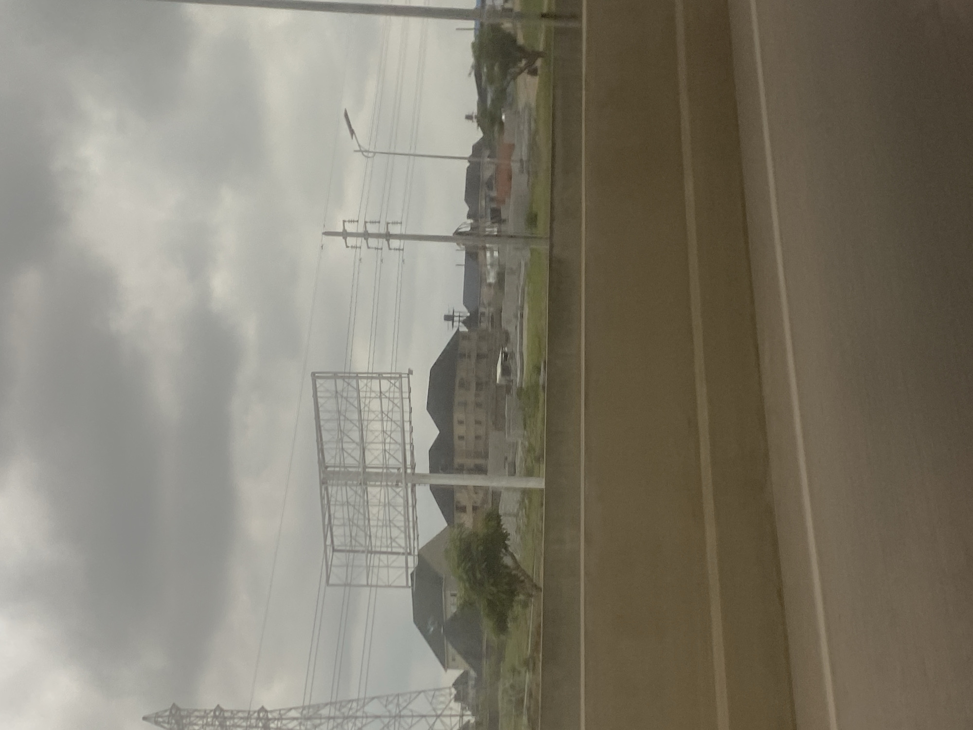 The image, oriented horizontally, depicts an outdoor scene observed from a perspective that suggests looking out from a vehicle or behind a fixed structure.
In the right foreground, a broad, light-colored surface with prominent vertical lines runs vertically through the frame, possibly representing a road, a highway barrier, or a part of the immediate viewing enclosure.
The left and central portions of the midground reveal an urban or suburban landscape. Numerous utility poles with extensive power lines crisscross the area, one featuring a streetlight. A large, skeletal metal billboard frame stands conspicuously in the mid-left, currently empty without any advertisements. Below and beyond these structures, several multi-story buildings with dark, sloped roofs are visible, some appearing residential or commercial. Sparse green trees and patchy vegetation dot the landscape between the buildings and beneath the power lines. The ground appears to be a mix of undeveloped earth and grass.
The background is dominated by an extensive, uniformly light grey and heavily overcast sky, indicating a gloomy or cloudy daytime.
No specific activities or people are discernible in the scene. The weather is characterized by heavy cloud cover. There is no visible text. The environment suggests an area undergoing development or an established infrastructure-heavy zone, consistent with a location like Gosa, Nigeria.