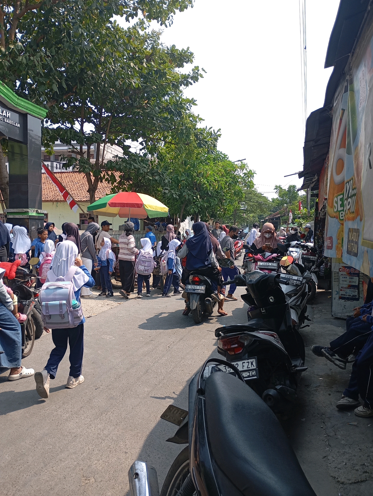 The image depicts a street scene in Babelan, Indonesia, likely near a school, given the presence of numerous children in school uniforms carrying backpacks. The street is paved and appears to be lined with trees and buildings. Several motorcycles are parked along the side of the road, some of which are occupied by people. A colorful umbrella stands in the middle of the street, providing shade. People are walking and standing, some engaged in conversation. The overall atmosphere suggests a busy morning or afternoon, possibly during a school commute.