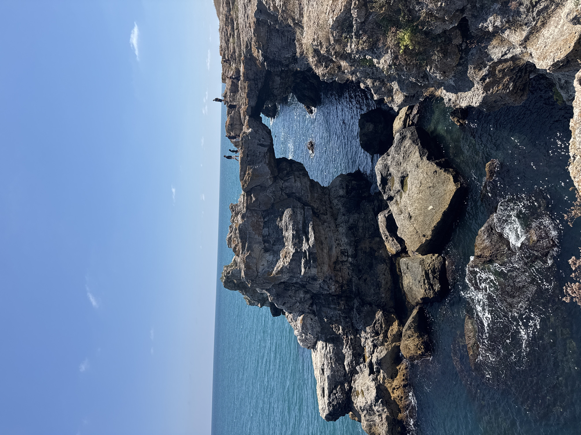 This image shows the natural rock arch formation known as The Arch in Tyulenovo, Bulgaria. The arch is a prominent geological feature carved by the sea from the rocky coastline. Several people are visible standing on the edge of the cliff and on top of the arch, observing the scenery and the sea. The sea is a deep blue, with some whitecaps and waves crashing against the rocks. The sky is clear and blue with a few scattered clouds. The overall environment is a rugged, natural coastal landscape.