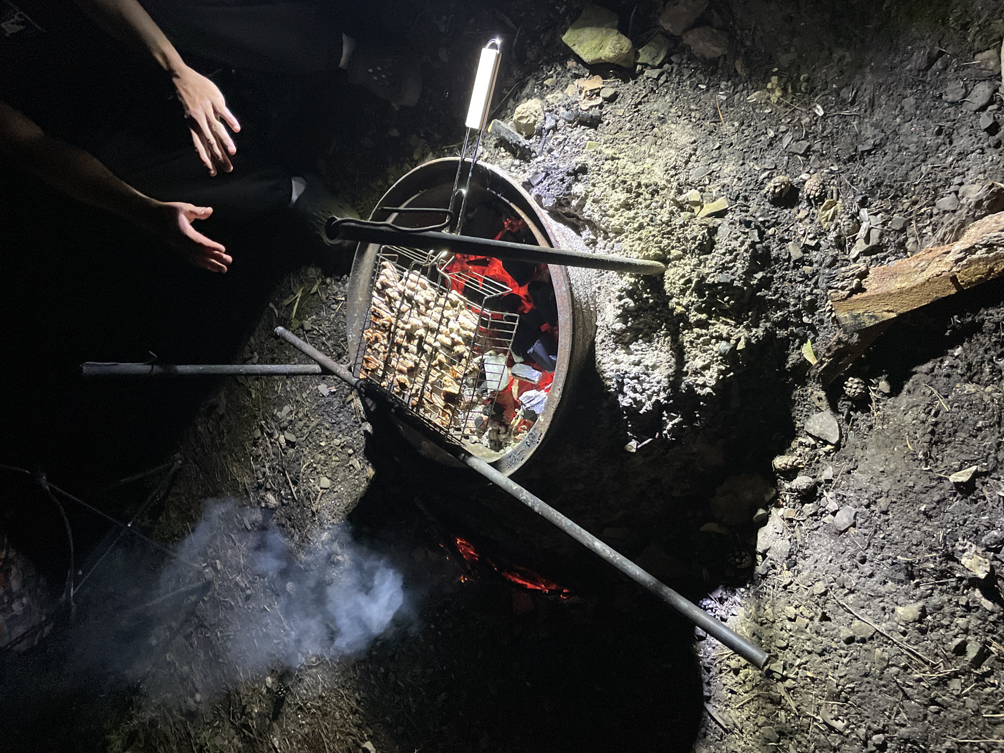 In the city of Nikolayevka, Russia, at night, a campfire is burning in a fire pit. A metal grill filled with food is suspended over the fire. Two people are present, with their hands visible near the grill, suggesting they are tending to the food. Smoke rises from the fire, illuminating the surrounding area with a warm glow. The ground is covered with dirt, rocks, and debris, indicative of an outdoor setting.
