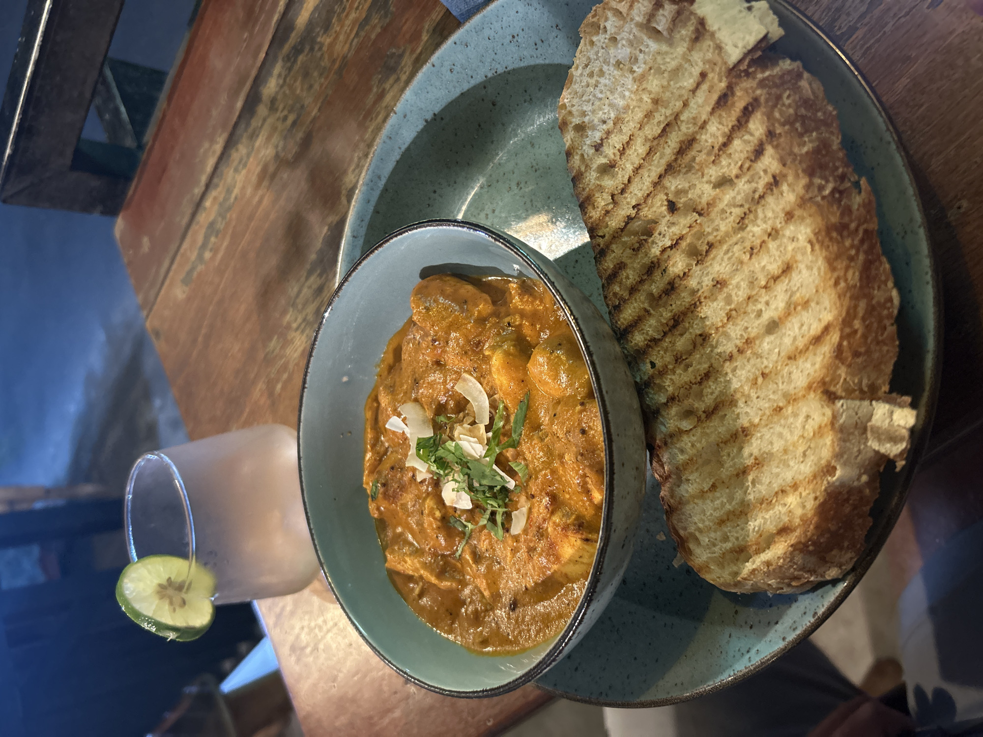 A close-up, overhead shot showcases a meal served on a rustic wooden table. The main focus is a blue-gray ceramic bowl filled with a vibrant, orange-hued curry, garnished with fresh green herbs and white flakes, likely coconut. Beside the bowl sits a piece of toasted bread with distinct grill marks, also on the same plate. To the left, a clear glass containing a pale, cloudy drink is visible, adorned with a slice of lime on the rim. The scene is set in what appears to be a restaurant or cafe, with hints of other tables or furniture in the background. The lighting suggests it could be daytime or evening, creating a warm and inviting atmosphere. The overall presentation of the food, along with the natural textures of the table and tableware, evokes a sense of casual dining and comfort. There is no visible text in the image. The setting appears to be the Ubud District in Indonesia.