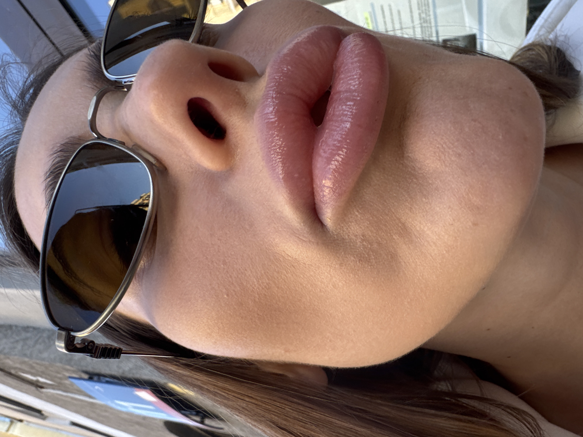 A close-up, upward-angle photograph captures a woman's face wearing sunglasses. Focus is on the lips, appearing full and glossy, situated above the nose. Parts of the sunglasses are visible, reflecting a bright, indistinct cityscape in the lenses. The woman's skin tone is fair. The background includes blurry details hinting at an outdoor environment, possibly in a city, with glimpses of other objects. Location specifics beyond the city of Warsaw are not clearly defined in the frame. The scene lacks definitive indications of actions or interactions.