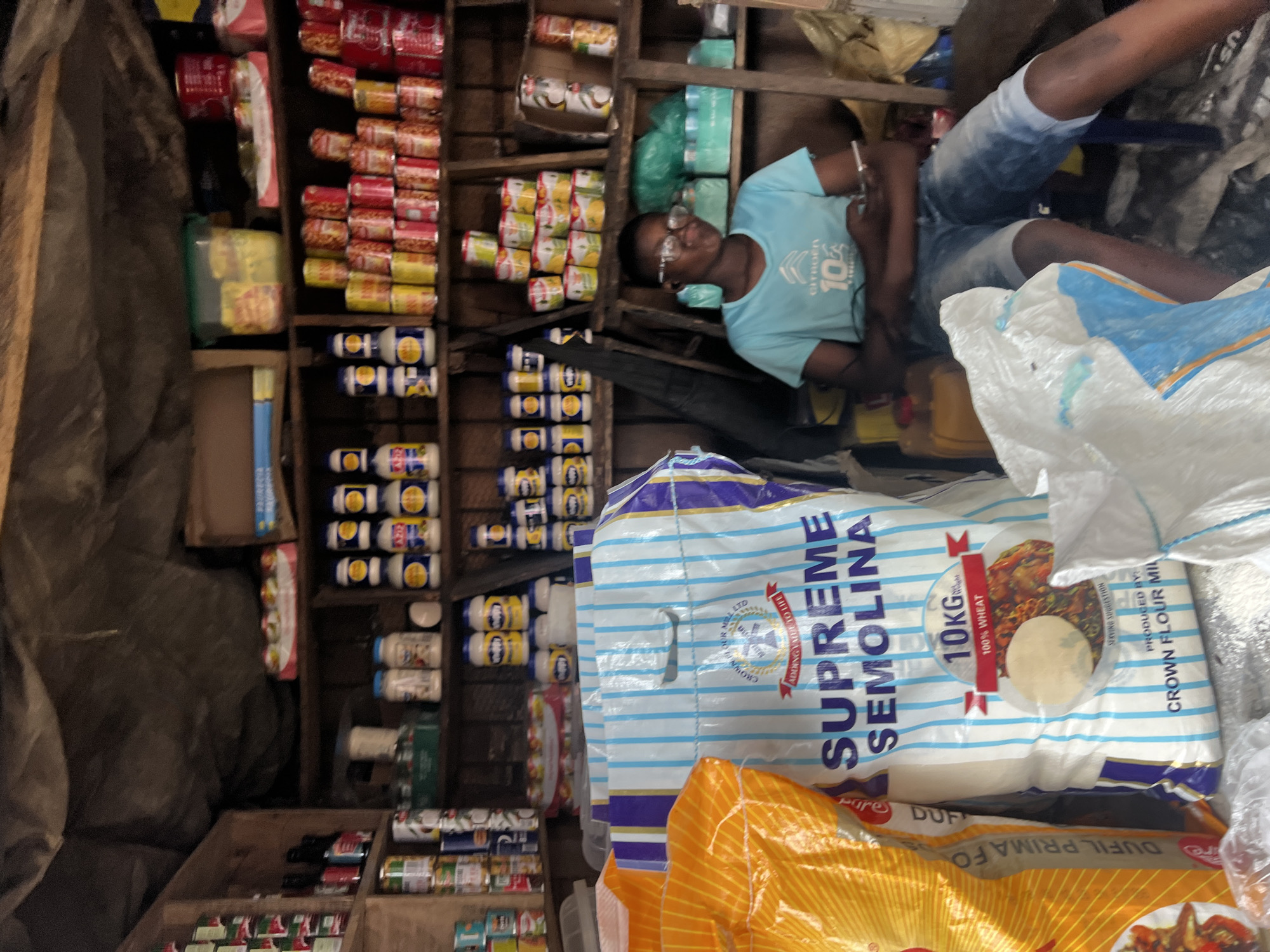 In a dimly lit market stall in Nkpor, Nigeria, a young person wearing glasses and a light blue t-shirt sits on a stool. The stall is stocked with various items, including bags of flour, canned goods, and bottles. A large bag labeled "SUPREME SEMOLINA 10KG 100% WHEAT" is prominently displayed in the foreground. The person is holding a smartphone and appears to be engaged with it. The overall impression is one of a busy, local market environment.