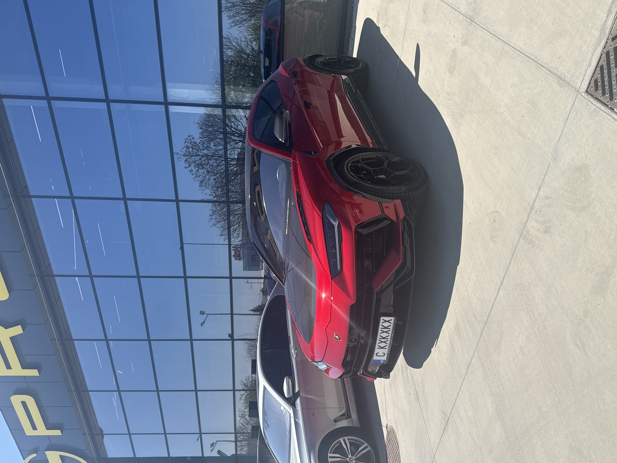 A red Lamborghini Urus SUV is prominently parked outdoors, facing the viewer, against a modern building with expansive reflective glass windows. The car is positioned on light-colored concrete pavement, which extends into the foreground and background. To the left of the Urus, partially visible, is a silver or light grey sedan.
The scene depicts a car dealership or a luxury showroom environment during bright daylight hours. Strong shadows cast on the pavement by the cars and an unseen object or person indicate direct sunlight. The sky, reflected in the large glass panels of the building, is clear and blue. Reflections of bare trees are also visible in the windows, suggesting a season like late winter or early spring.
Notable details include the aggressive styling of the red Lamborghini Urus, featuring black accents and black multi-spoke wheels with "SCORPION" visible on the tire sidewalls. A metal grate is present in the concrete pavement in the bottom right corner. On the building facade, large yellow letters "P R" (or "P O R") are partially visible on the left side of the image, possibly indicating a brand or dealership name. The license plate on the front of the Lamborghini Urus reads "C KXXXXX", with a small blue strip on the left displaying "BG" and "STUDION", confirming it is a Bulgarian license plate. The overall impression is one of luxury vehicles presented in a pristine, contemporary setting in Plovdiv, Bulgaria. No people or overt activity are visible in the image.