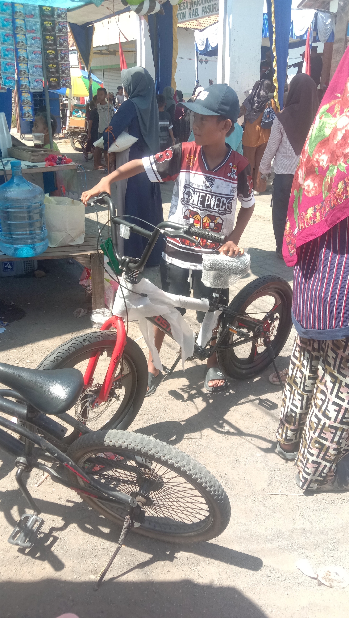The image depicts an outdoor scene, likely a marketplace or a gathering, in the city of Karang Anyar, Indonesia. The foreground features two bicycles. One bicycle, predominantly black with red accents on its front forks and wheel rims, is partially visible on the left. The second bicycle, wrapped in white plastic sheeting, is positioned centrally. A young boy wearing a dark baseball cap and a white t-shirt with "ONE PIECE" graphics and black and red sleeves is standing next to this bicycle, his right hand on the handlebars. Behind him, other individuals are present, including a woman wearing a dark hijab and a patterned skirt, and another woman in a striped top and patterned pants. A large blue water dispenser is visible on a wooden table to the left. In the background, tents, stalls, and more people can be seen, suggesting a lively event. The ground is dirt or sand.