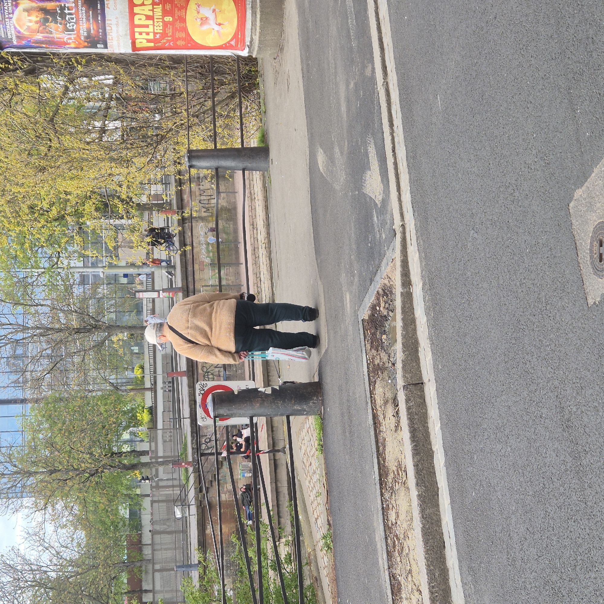The image depicts a street scene in Strasbourg, France. An elderly person wearing a beige jacket, dark pants, and a white hat is standing on the sidewalk. The individual carries a white bag. The sidewalk is adjacent to a street and a fenced area with trees. A billboard displays advertisements, including one for "Alsati," an event in Strasbourg, and another for the "Pelpass Festival." The surrounding environment includes buildings and foliage.