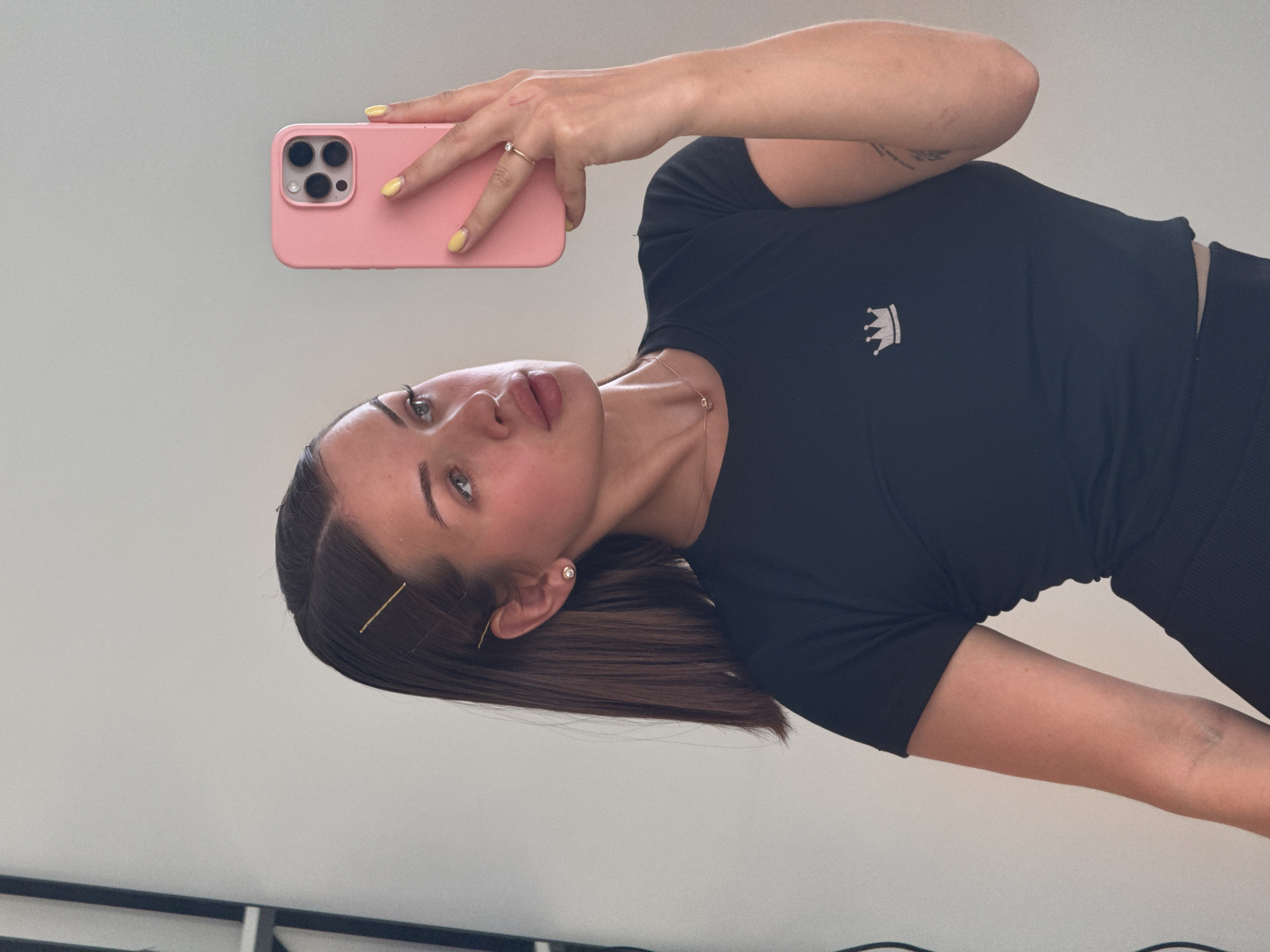 A young woman with brown hair styled with bobby pins is taking a selfie in a mirror. She is wearing a black t-shirt with a crown logo and dark leggings. She has on a thin gold necklace with a small pendant, and a pearl earring. She is also wearing a gold ring on her right hand. The woman's phone, which is in a pink case, is visible as she holds it up to take the photo. The background is a plain, light-colored wall. The image does not contain any information about the city of Perm, Russia.