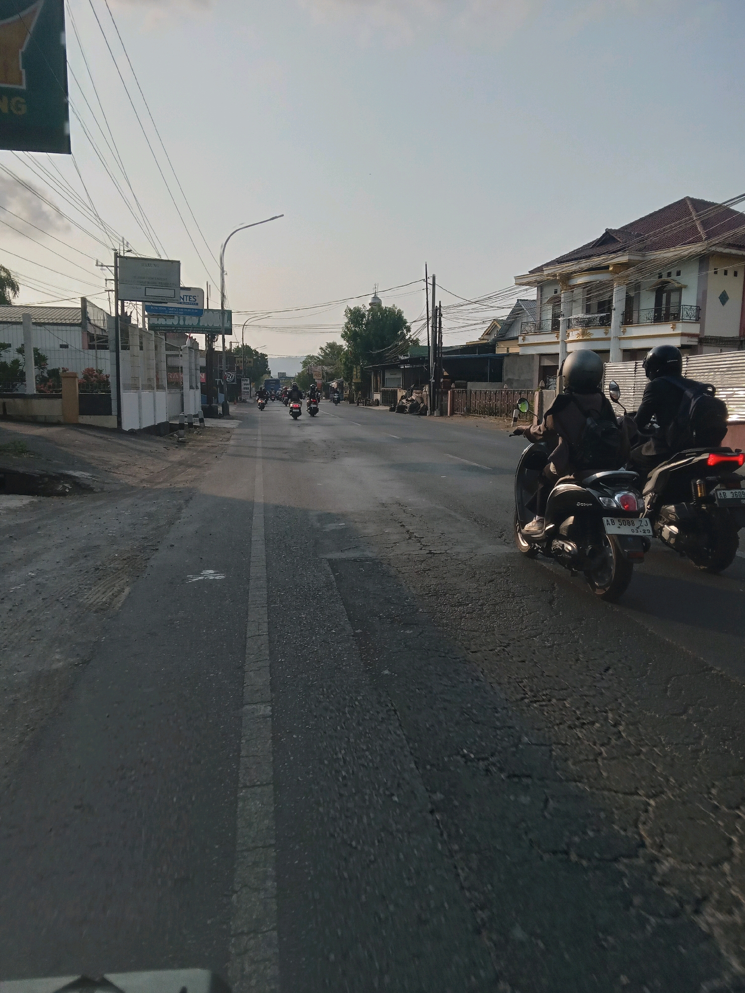 The image shows a street scene in Berbah, Indonesia, likely during daytime with clear skies. Two individuals are riding on separate scooters, one in front of the other, on the right side of the paved road. Both riders are wearing helmets and backpacks. The scooter in the foreground has a license plate reading "AB 5088 ZJ" and "03.25". The scooter behind it has a license plate reading "AB 3609 JXL". The road surface is asphalt and appears cracked in some areas. On the left side of the road, there are buildings with various signs, including one with text that seems to be in Indonesian. Overhead power lines are visible crisscrossing the sky. In the background, more buildings and trees are present, with a distant view of hills or mountains. The overall impression is a typical urban or suburban street in Indonesia.