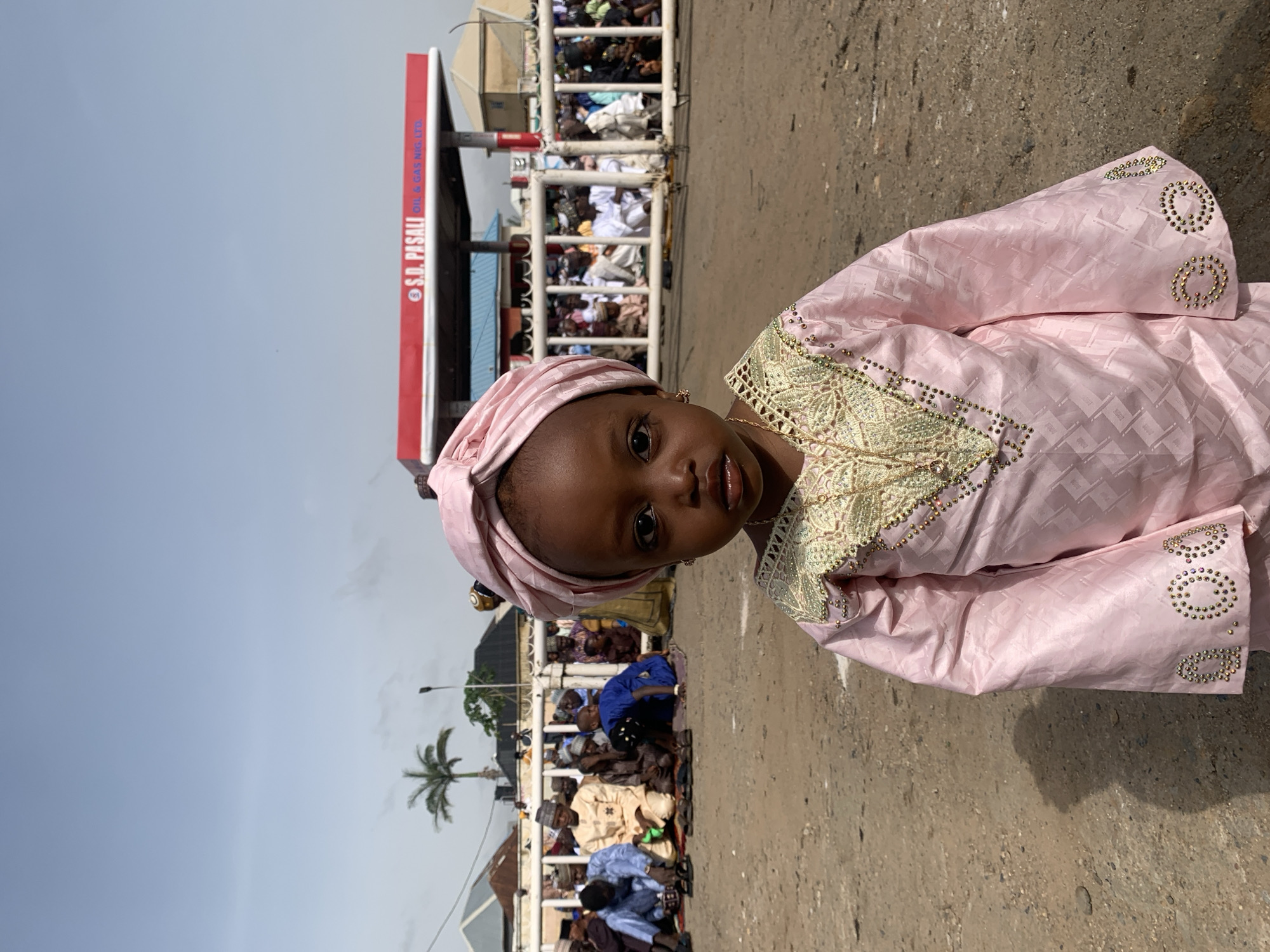 A young child with dark skin and brown eyes is centered and facing forward, dressed in a pink traditional garment and headwrap with gold embellishments. The photo seems to be taken in Jos, Nigeria, given the presence of an S.D. PASALI Oil & Gas Nig. LTD. gas station in the background, visible behind a white metal barricade. Behind the barricade are numerous people gathered, possibly praying or participating in an event. Some individuals are kneeling or sitting on the ground. The environment includes a palm tree, a streetlamp, buildings, and overcast skies. The surface the child is standing on appears to be a gray, concrete road or sidewalk.