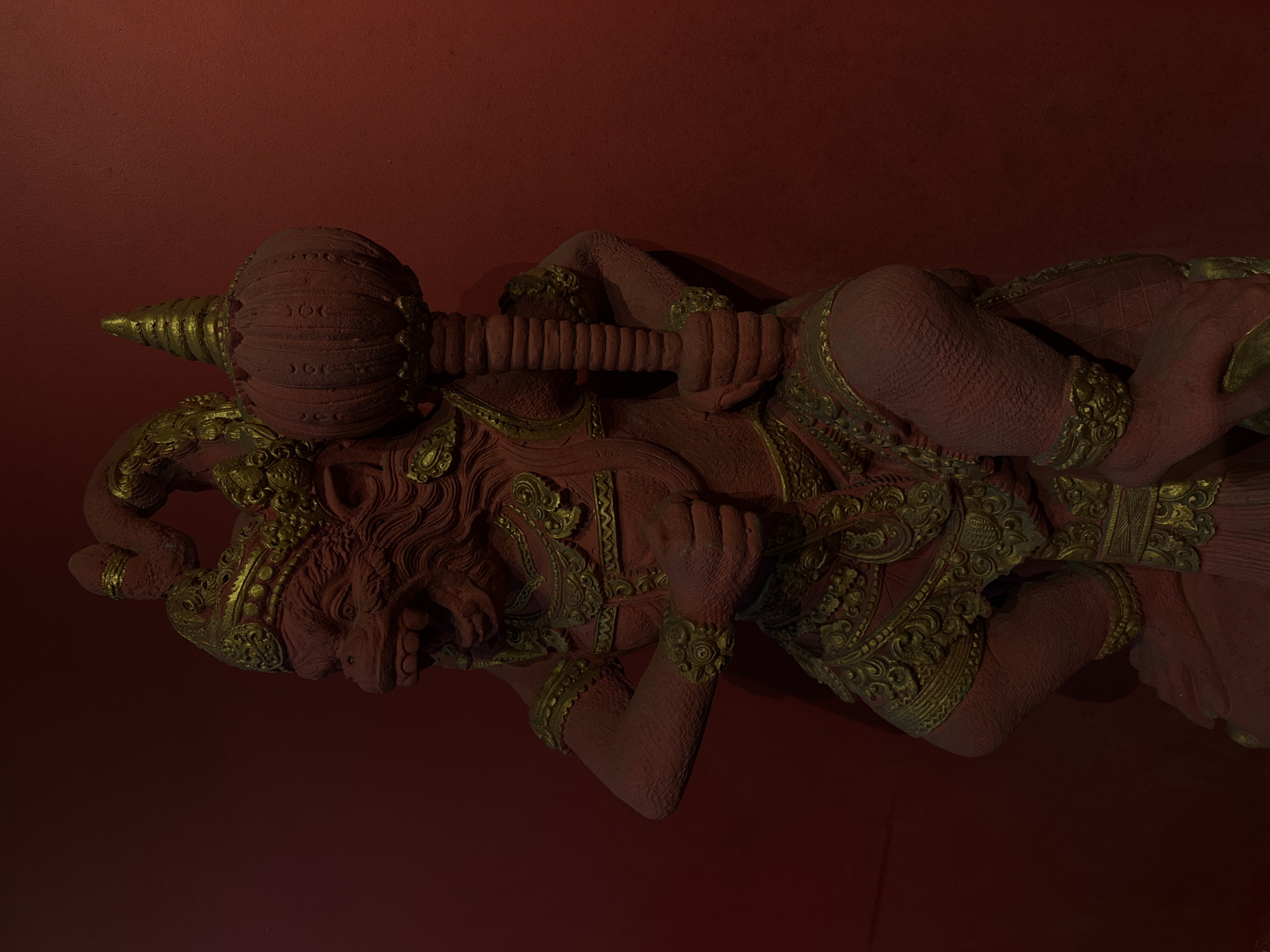 The image displays a close-up, overhead view of a large, red, stone statue with gold accents, resting against a deep red wall. The statue depicts a mythical creature or deity, possibly a demon or a warrior, with a fierce expression, sharp teeth, and a mane-like hairstyle. It is adorned with ornate gold embellishments on its headdress, shoulders, chest, arms, and legs, suggesting a rich cultural or religious significance.
The creature is depicted in a reclining or resting pose, holding a large club-like object horizontally across its body. The club itself is textured and also features gold detailing. The statue has a rough, textured surface, indicating it is likely carved from stone or a similar material. The lighting is dramatic, with strong shadows obscuring parts of the statue and highlighting its form and intricate details. The background is a plain, textured red wall, which creates a stark contrast with the statue. There is no visible text in the image. The overall mood is one of power and ancient grandeur, amplified by the dramatic lighting and deep colors. Given the location context of Kuta, Indonesia, this statue could be part of a temple, a traditional performance stage, or a decorative element in a public space, reflecting Balinese or Indonesian artistic and mythological traditions. The time of day is not apparent, but the lighting suggests it might be indoors or under artificial illumination.
