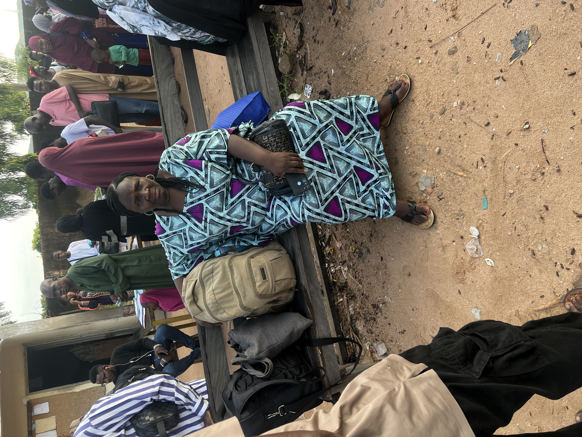 The image depicts an outdoor public gathering in Gbaza, Nigeria, centered around a wooden bench on a reddish-brown sandy ground. A woman of color, the primary subject, is seated on the bench, facing forward with a neutral expression. She wears a long V-neck dress adorned with a geometric pattern of light blue, white, and purple triangles, black flip-flops with brown soles, and red nail polish on her toes. In her left hand, she holds a small black textured purse and a black phone or wallet.
To her right on the bench, a large tan canvas backpack, a gray fabric tote bag, and a black padded backpack are placed. A blue bag or fabric is visible behind her to her left. The sandy ground in the foreground and around the bench is littered with small pieces of plastic debris, paper, bottle caps, and organic matter.
In the bottom-left foreground, a person is partially visible, wearing a tan short-sleeved shirt and black cargo pants, with brown sandals on the ground.
The mid-ground and background are populated by a diverse crowd. On the left, a man in a black long-sleeved shirt and glasses is seated on a stool, wearing earphones, near a light-colored building with a door and papers affixed to its exterior wall. Adjacent to him, a balding man in a long green robe stands, holding a small object. Behind them, a woman in an orange and black leopard print top is visible.
On the right side of the background, a dense group of people stands. This group includes men in various attire such as a pink polo shirt and a tan long robe, and several women wearing head coverings (hijabs) in colors like maroon and black. A small child in a green shirt is visible among the crowd. The background also features a light-colored wall or fence and distant green foliage under an overcast sky. The scene suggests a busy communal waiting or gathering area.