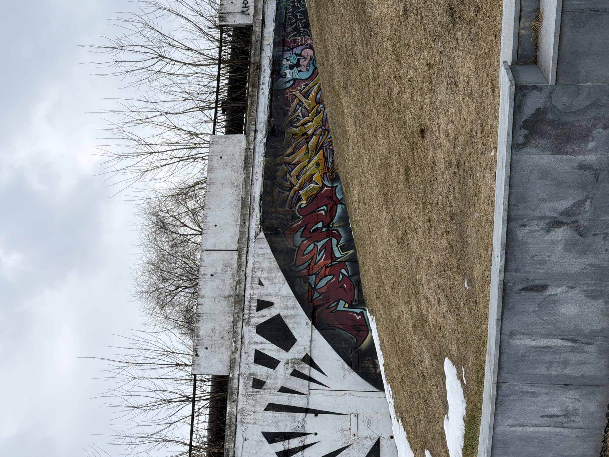 The image depicts a concrete retaining wall in Votkinsk, Russia, adorned with graffiti art. The wall features both geometric black and white patterns and colorful spray-painted designs, some of which are cartoonish and some appear to be lettering. A grassy slope rises to the right of the wall, showing patches of snow. Bare tree branches are visible against a gray, overcast sky above the wall. A section of a paved area, possibly a walkway or road, is partially visible in the lower right corner. There are no people visible in the photograph.