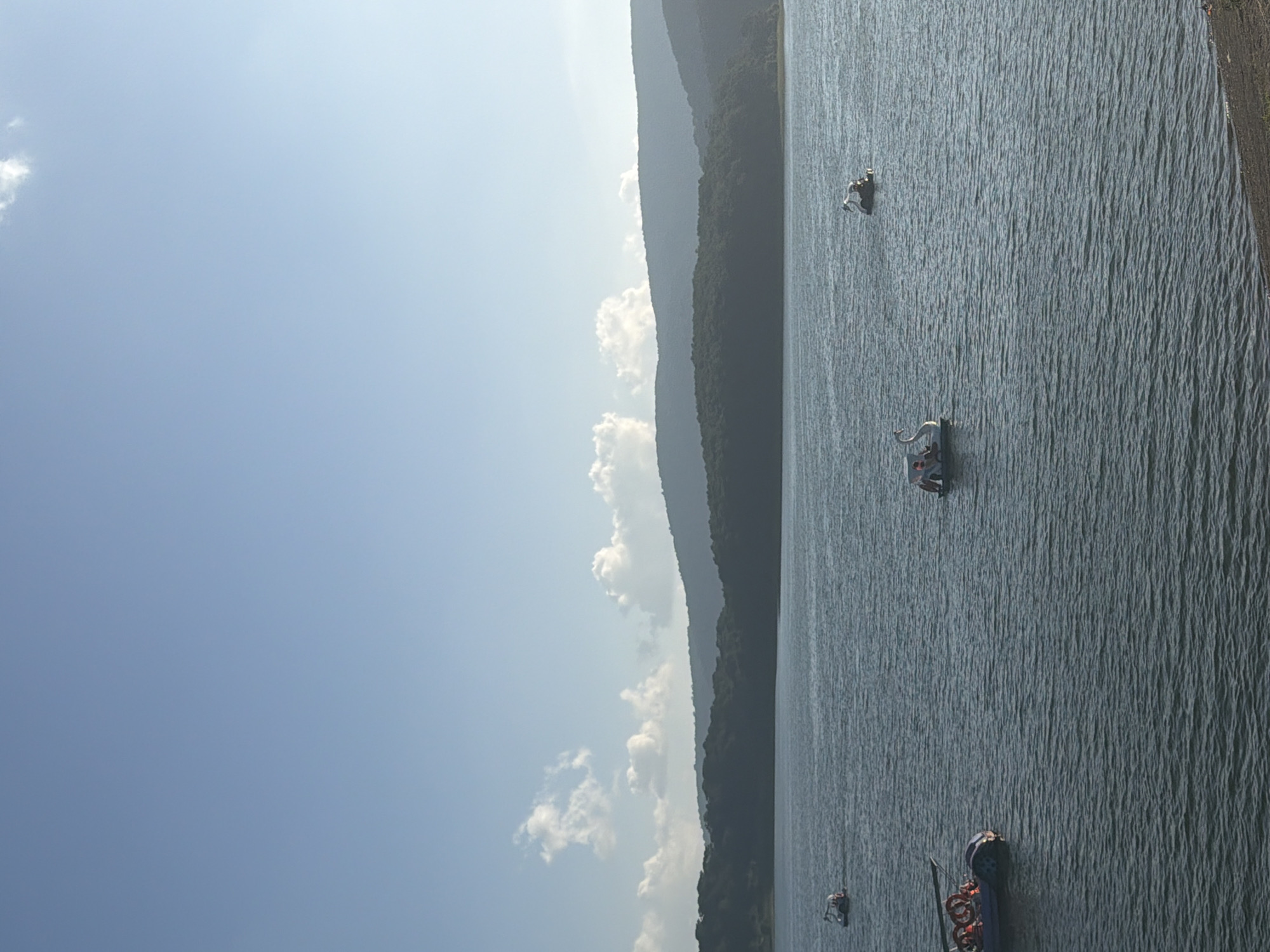 The image depicts a tranquil scene on a large body of water, likely a lake or reservoir, situated in the city of Da Lat, Vietnam. The foreground is dominated by the dark, rippling surface of the water, with a small strip of sandy shore visible at the bottom right. Several recreational boats are scattered across the water. On the left, a red and blue inflatable boat with life preservers and passengers is being steered. Further out, two swan-shaped pedal boats are visible, each carrying people. Another small boat is seen in the distance on the left. In the mid-ground, a dark, tree-covered hillside forms a dense band, suggesting a forested area. Beyond this, a series of layered, hazy blue mountains recede into the distance, creating a sense of depth and vastness. The sky above is a clear, pale blue, with a few wispy white clouds scattered across the upper portion of the frame. The overall impression is one of a peaceful, outdoor recreational setting on a clear day.