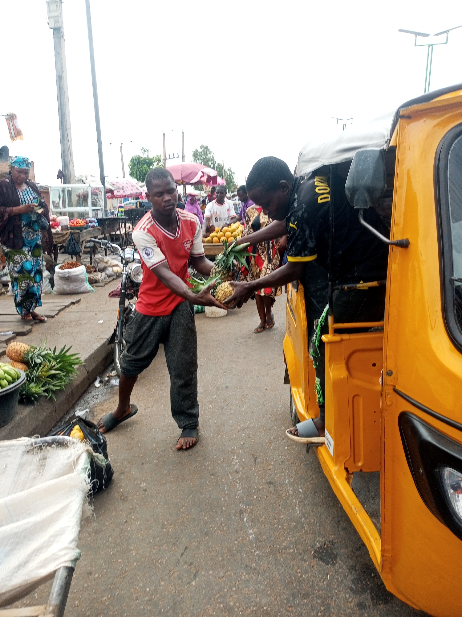 The image depicts a market scene in Tempure, Nigeria. Two men are in the foreground. One, wearing a red Arsenal jersey and gray pants, stands outside a yellow auto-rickshaw (tuk-tuk) and is handing pineapples to the other man, who is inside the vehicle. Other people and market stalls are visible in the background, suggesting a busy outdoor marketplace. There is a bicycle parked behind the man in the red jersey. Other objects visible include bags of produce, other fruits, and some umbrellas. The environment appears to be an open market area with a concrete curb and paved road.