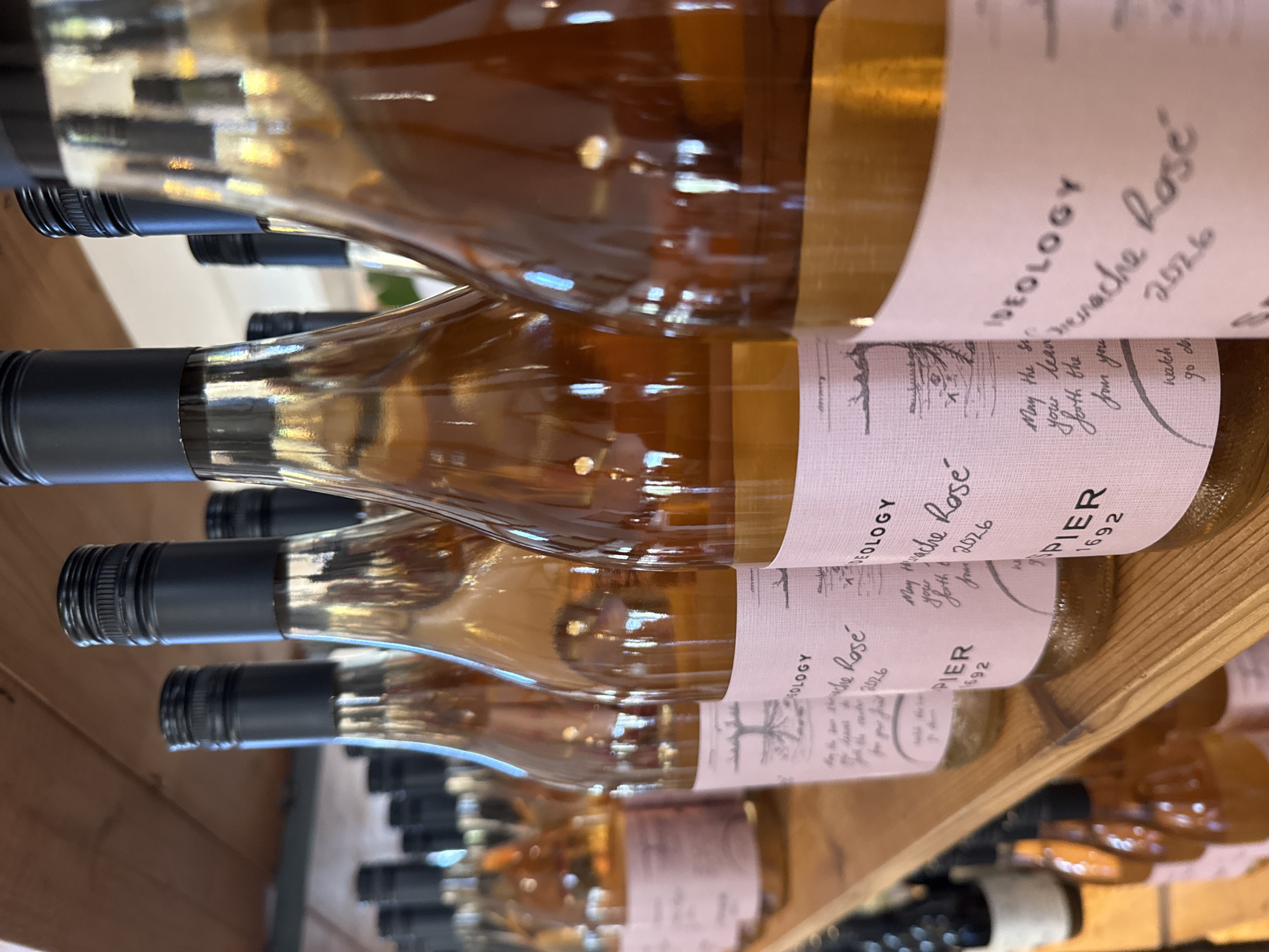 This image depicts a close-up view of several bottles of rosé wine lined up on a wooden shelf. The bottles have light pink labels with text and illustrations. The text on the visible labels includes "IDEOLOGY", "Grenache Rosé", a date of "2026", and "PIER 1692". Some labels also feature a drawing of a tree with roots. The wine inside the bottles appears to be a light amber or pinkish-orange color. The shelves are made of light-colored wood, and the background shows more bottles and a wooden wall, suggesting a wine cellar, store, or tasting room. The lighting is warm and highlights the reflections on the glass bottles. This scene is likely located within a wine establishment in Stellenbosch Nu, South Africa, given the prominent display of wine bottles and the commonality of wineries and wine-related businesses in that region. There are no people visible in the image.
